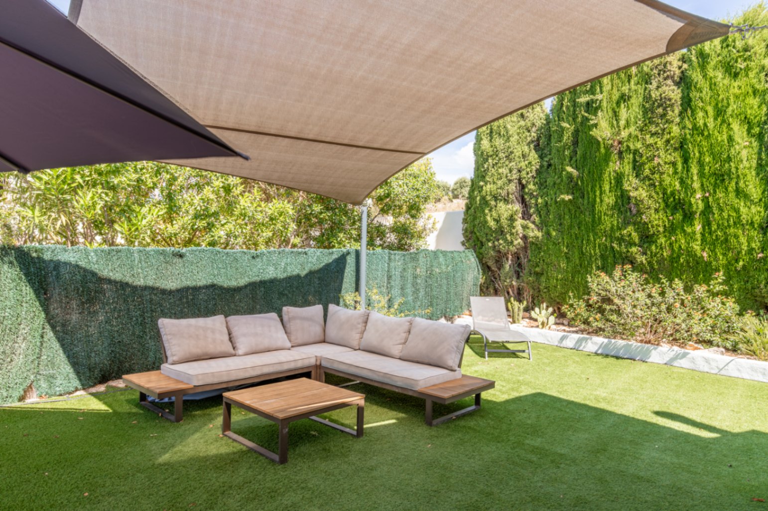 Schattiger Außenbereich mit L-förmigem Sofa, hellen Kissen und Holztisch auf Kunstrasen. Hohe Hecken und Bäume bieten Privatsphäre unter einem großen beigen Sonnensegel.