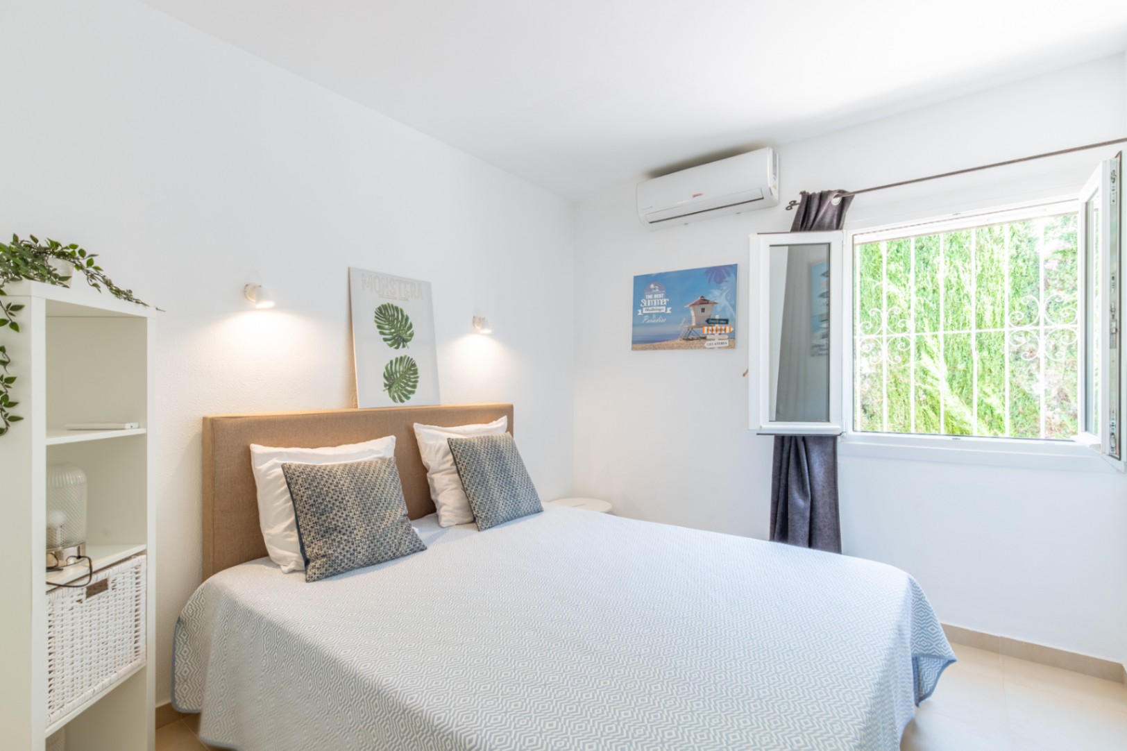 Schlafzimmer mit Doppelbett, beigem Stoffkopfteil, Klimaanlage und Fenster mit Sicherheitsgitter, das den Blick auf grüne Vegetation freigibt.