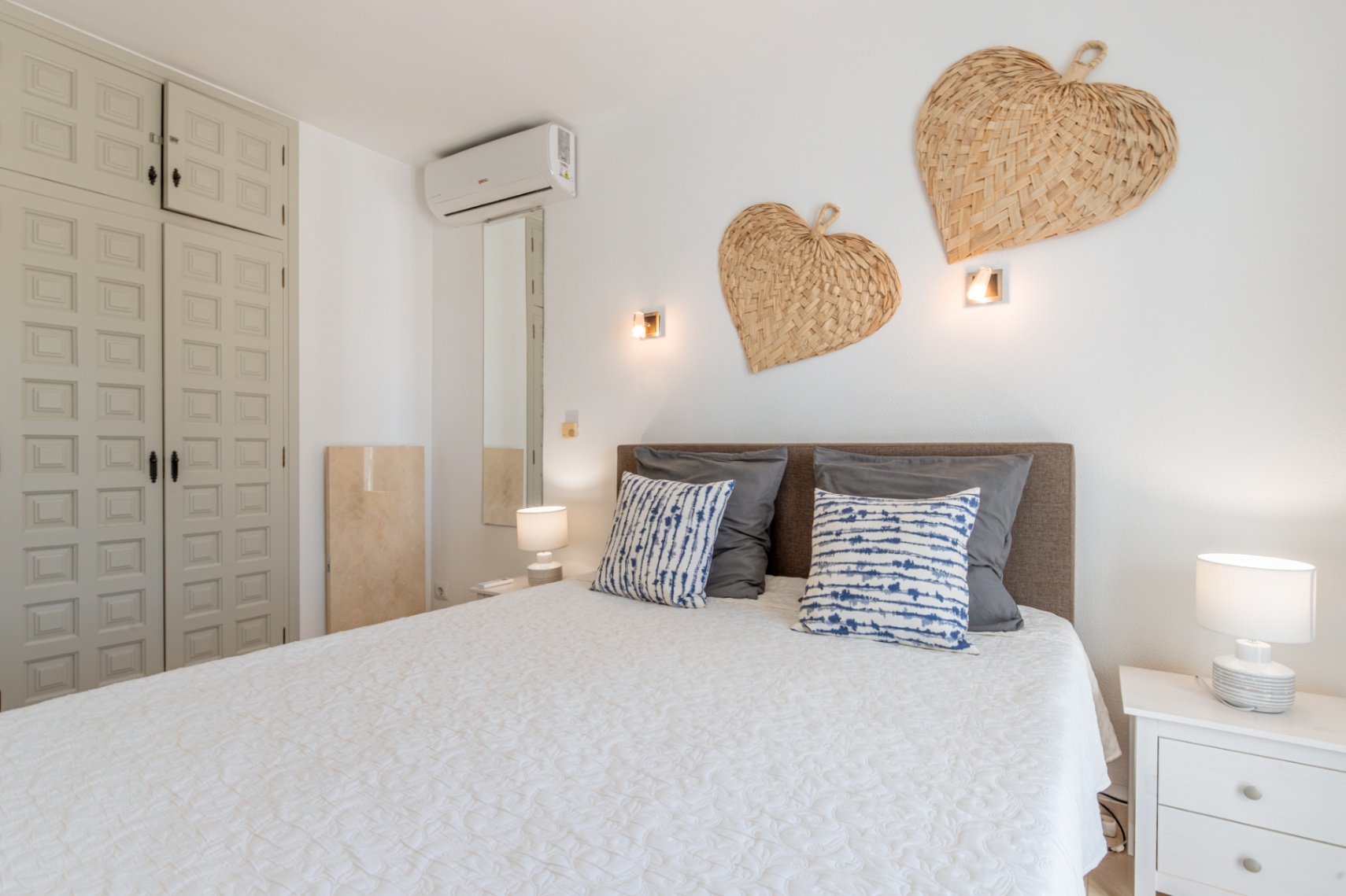 Schlafzimmer mit gemütlichem Bett, grauem Kopfteil, weißer Steppdecke und gemusterten Kissen. Einbauschrank mit Paneelen, Wandklimaanlage und geflochtenen Herzdekorationen.