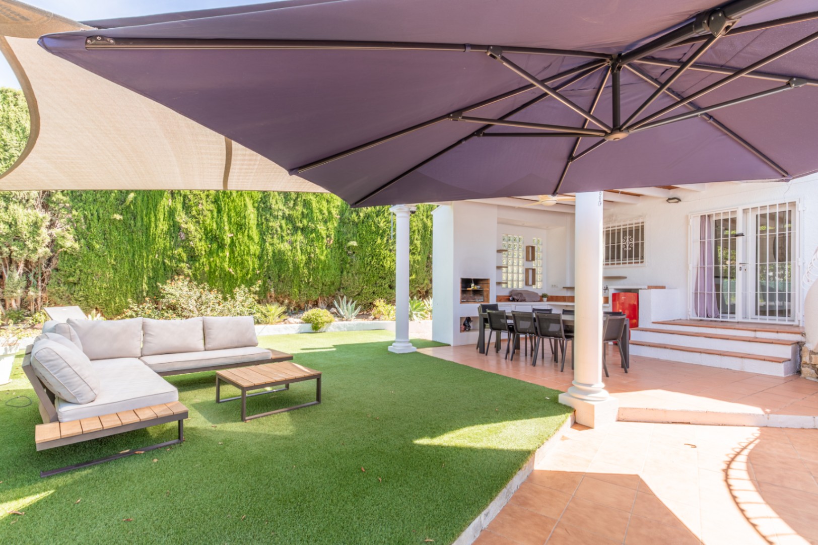 Überdachte Außenterrasse mit Essbereich und integriertem Grill, angrenzend an einen Kunstrasenbereich mit Lounge-Möbeln und großem Sonnenschirm.