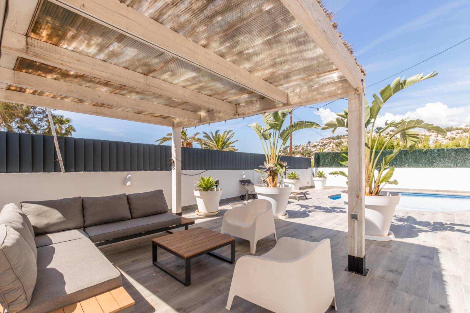 Überdachte Außenterrasse mit Holzpergola und transluzentem Dach, ausgestattet mit grauem Modulsofa, Couchtisch und tropischen Pflanzen. Grauer Fliesenboden mit teilweisem Poolblick.