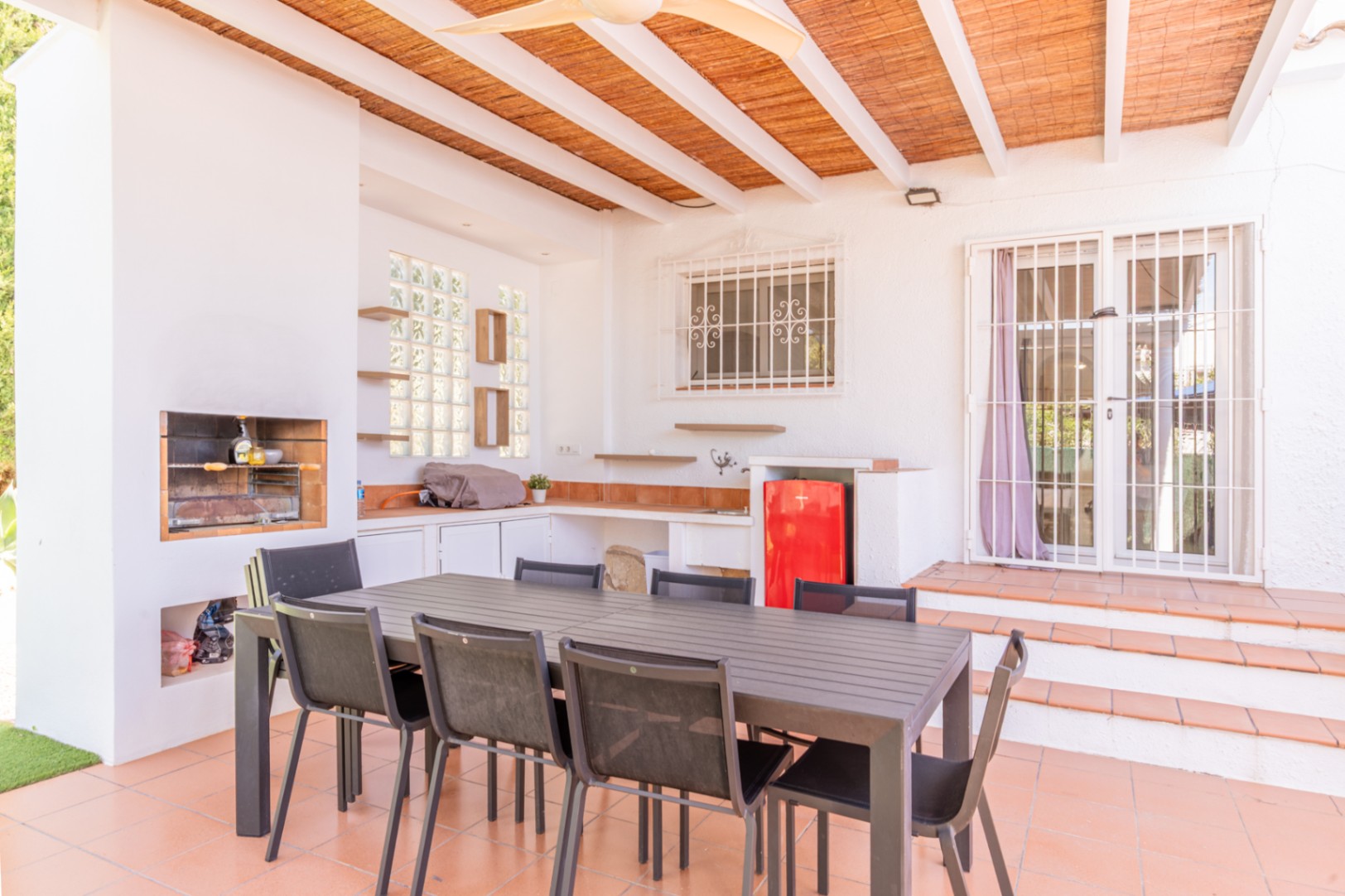 Überdachte Außenterrasse mit integriertem Grill, funktionaler Außenküche inklusive rotem Minikühlschrank und großem Esstisch. Der Bereich verfügt über Terrakottaboden und eine Lattenholzdecke.