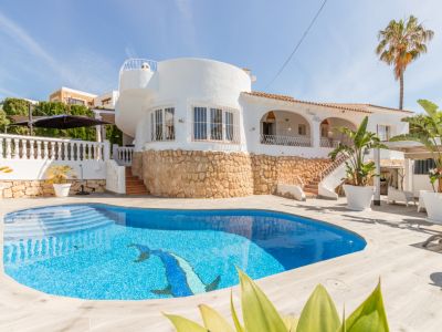 Außenansicht einer weißen Villa mit privatem blau gefliestem Pool und Delfinmosaik. Steinfassade und ein markanter runder Gebäudeteil sind sichtbar.