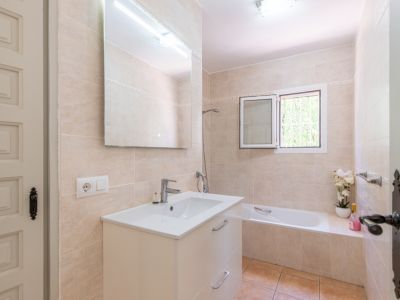 Badezimmer mit hellbeigen Fliesen, weißem Waschtischunterschrank und großem Spiegel mit Beleuchtung. Ausgestattet mit Badewanne und Fenster für Tageslicht.