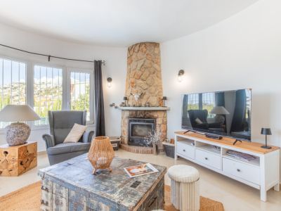 Gemütliches Wohnzimmer mit steingemauertem Kamin und großen Erkerfenstern mit Blick auf die Hügel. Ein rustikaler Couchtisch und ein modernes TV-Möbel ergänzen den Raum.