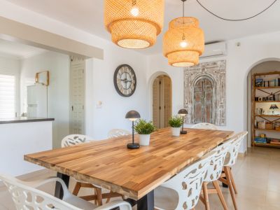 Geräumiger Essbereich mit Holztisch, modernen weißen Stühlen und zwei geflochtenen Hängelampen. Bögen führen zur Küche und einem Lesebereich mit Holzregal.