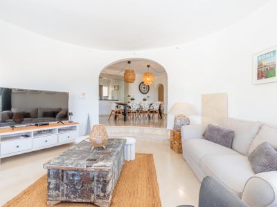 Geräumiges Wohnzimmer mit hellen Fliesen, bequemen Sofa und rustikalem Holztisch auf Jute-Teppich. Ein Bogen führt zum erhöhten Essbereich mit modernen Stühlen und Rattan-Hängeleuchten.