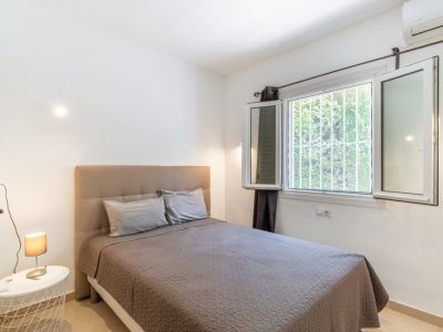 Helles Schlafzimmer mit bequemem Bett und gepolstertem Kopfteil, großem Fenster mit Sicherheitsgittern und Blick ins Grüne, sowie Klimaanlage.