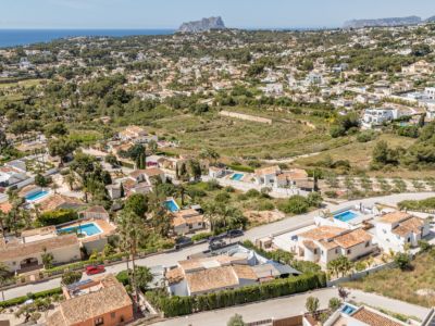 Luftaufnahme eines Wohngebiets in Moraira, mit zahlreichen Villen mit Terrakottadächern, privaten Pools und üppiger Vegetation. Das Mittelmeer und eine ferne Felsformation prägen die Küstenlandschaft.