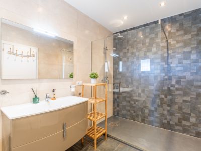 Modernes Badezimmer mit glänzend beigem Waschtisch, beleuchtetem Spiegel und ebenerdiger Dusche mit Glaswand und dunkelgrauen Mosaikfliesen. Helle Wandfliesen, dunkler Boden.