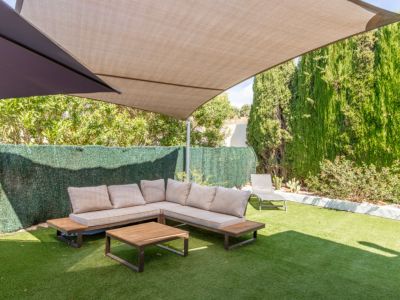 Schattiger Außenbereich mit L-förmigem Sofa, hellen Kissen und Holztisch auf Kunstrasen. Hohe Hecken und Bäume bieten Privatsphäre unter einem großen beigen Sonnensegel.