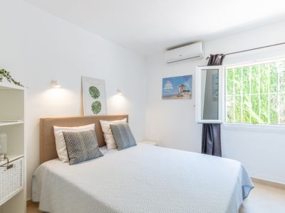 Schlafzimmer mit Doppelbett, beigem Stoffkopfteil, Klimaanlage und Fenster mit Sicherheitsgitter, das den Blick auf grüne Vegetation freigibt.