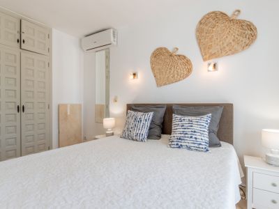 Schlafzimmer mit gemütlichem Bett, grauem Kopfteil, weißer Steppdecke und gemusterten Kissen. Einbauschrank mit Paneelen, Wandklimaanlage und geflochtenen Herzdekorationen.
