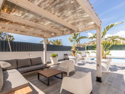 Überdachte Außenterrasse mit Holzpergola und transluzentem Dach, ausgestattet mit grauem Modulsofa, Couchtisch und tropischen Pflanzen. Grauer Fliesenboden mit teilweisem Poolblick.