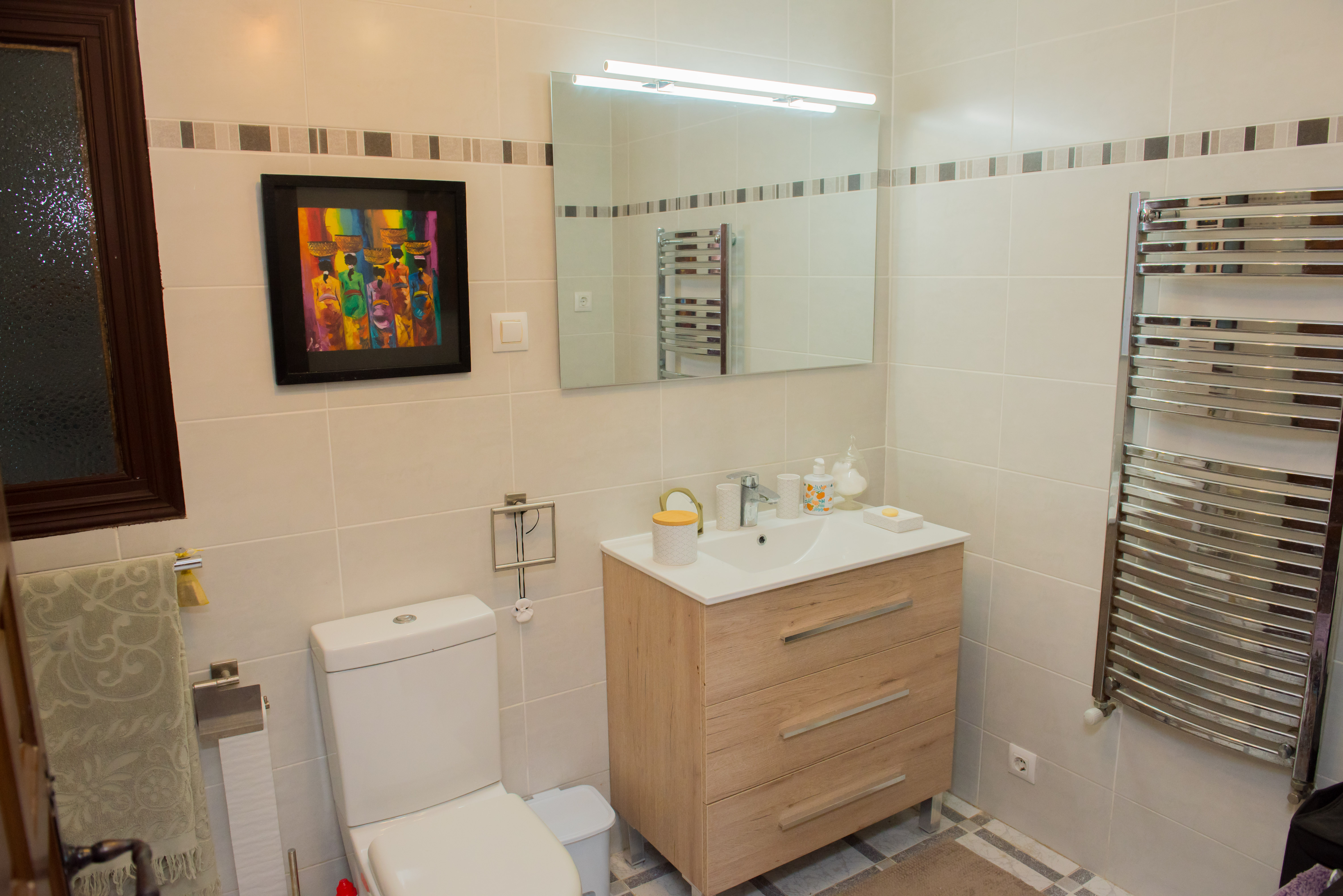 Badezimmer mit cremefarbenen Fliesen, hellem Holzwaschtisch und verchromtem Handtuchwärmer.
