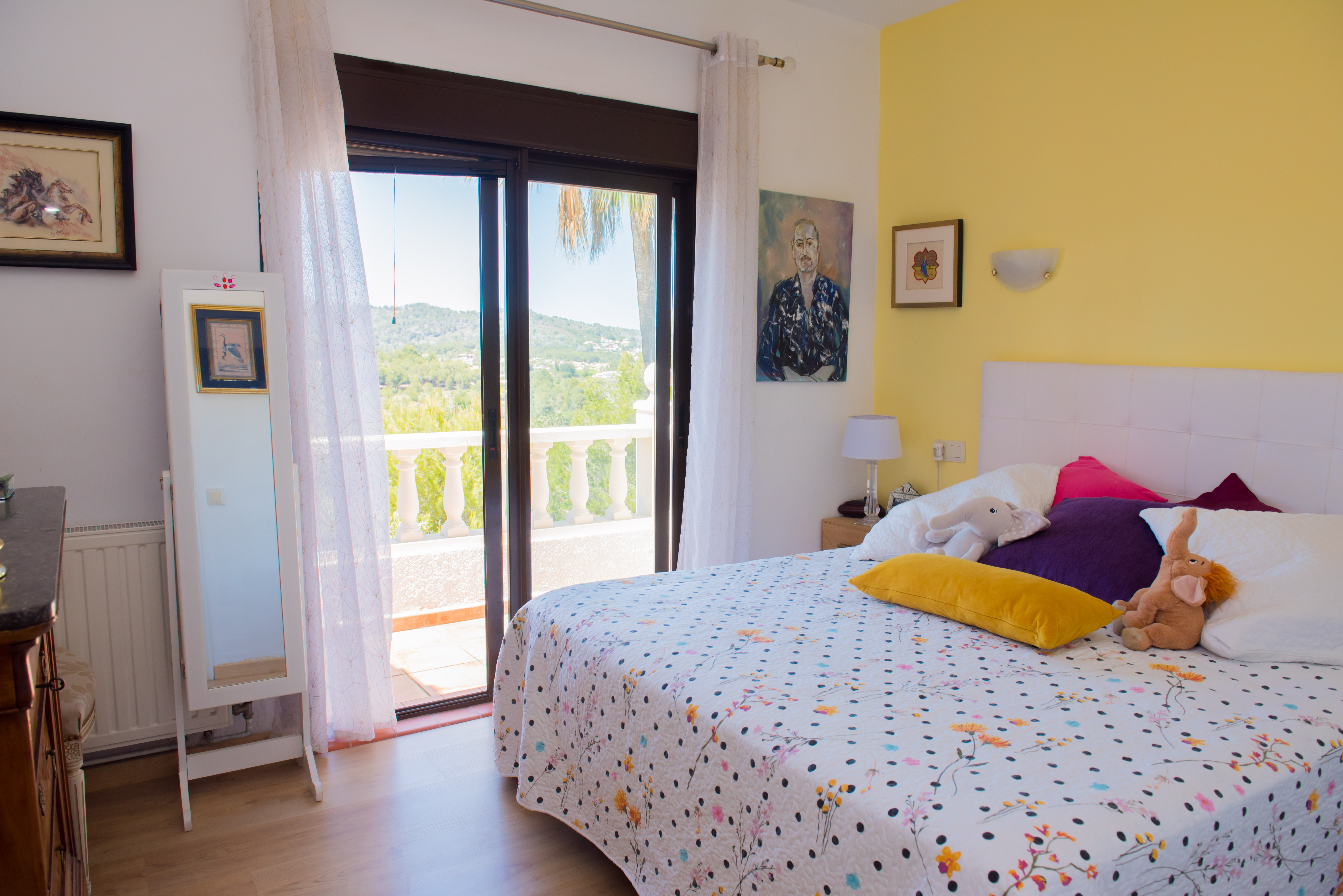 Helles Schlafzimmer mit Balkonzugang. Gelbe Akzentwand und gemusterte Bettwäsche.
