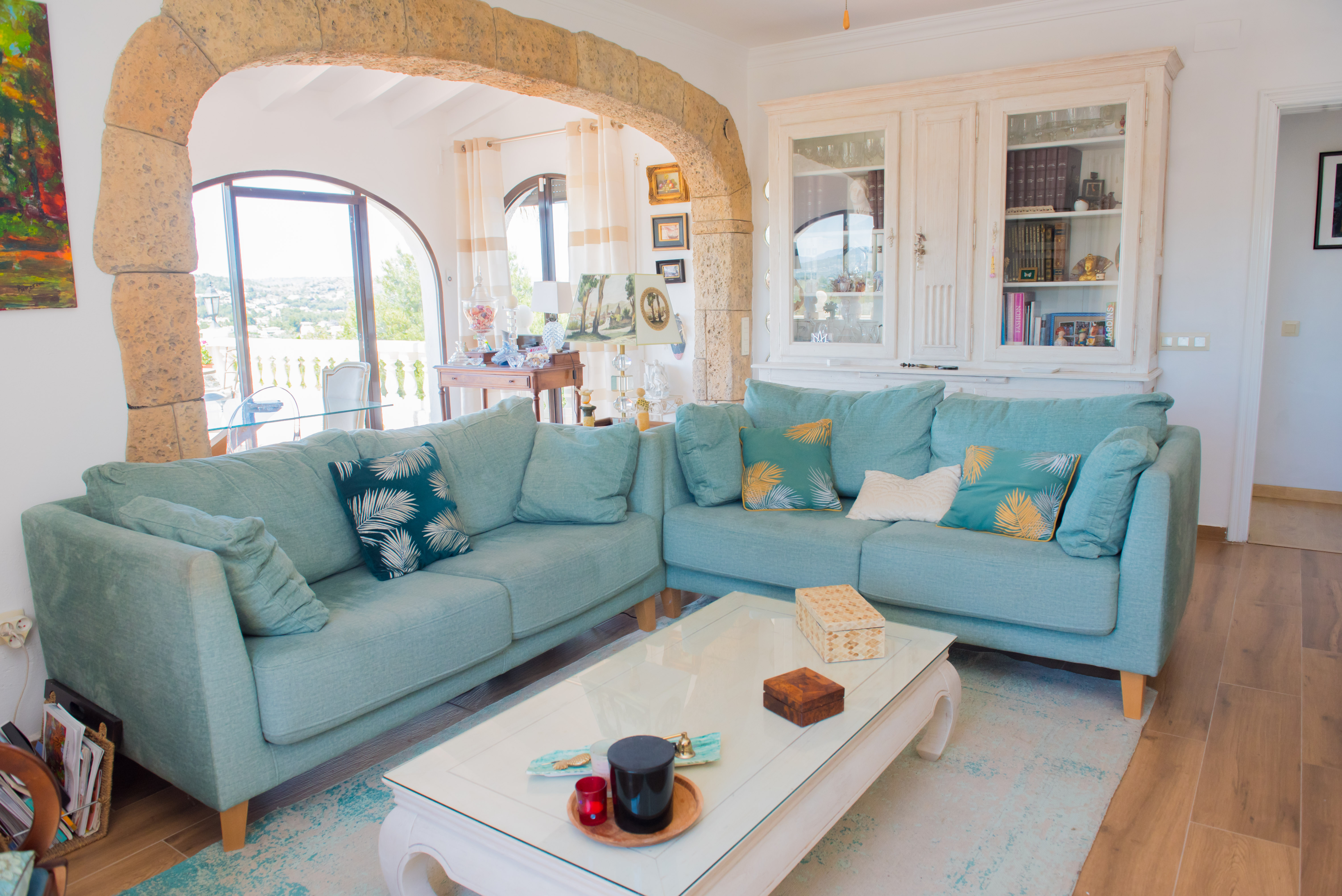 Helles Wohnzimmer mit türkisfarbenem Ecksofa, weißem Couchtisch und rustikalem Steinbogen mit Blick ins Freie.
