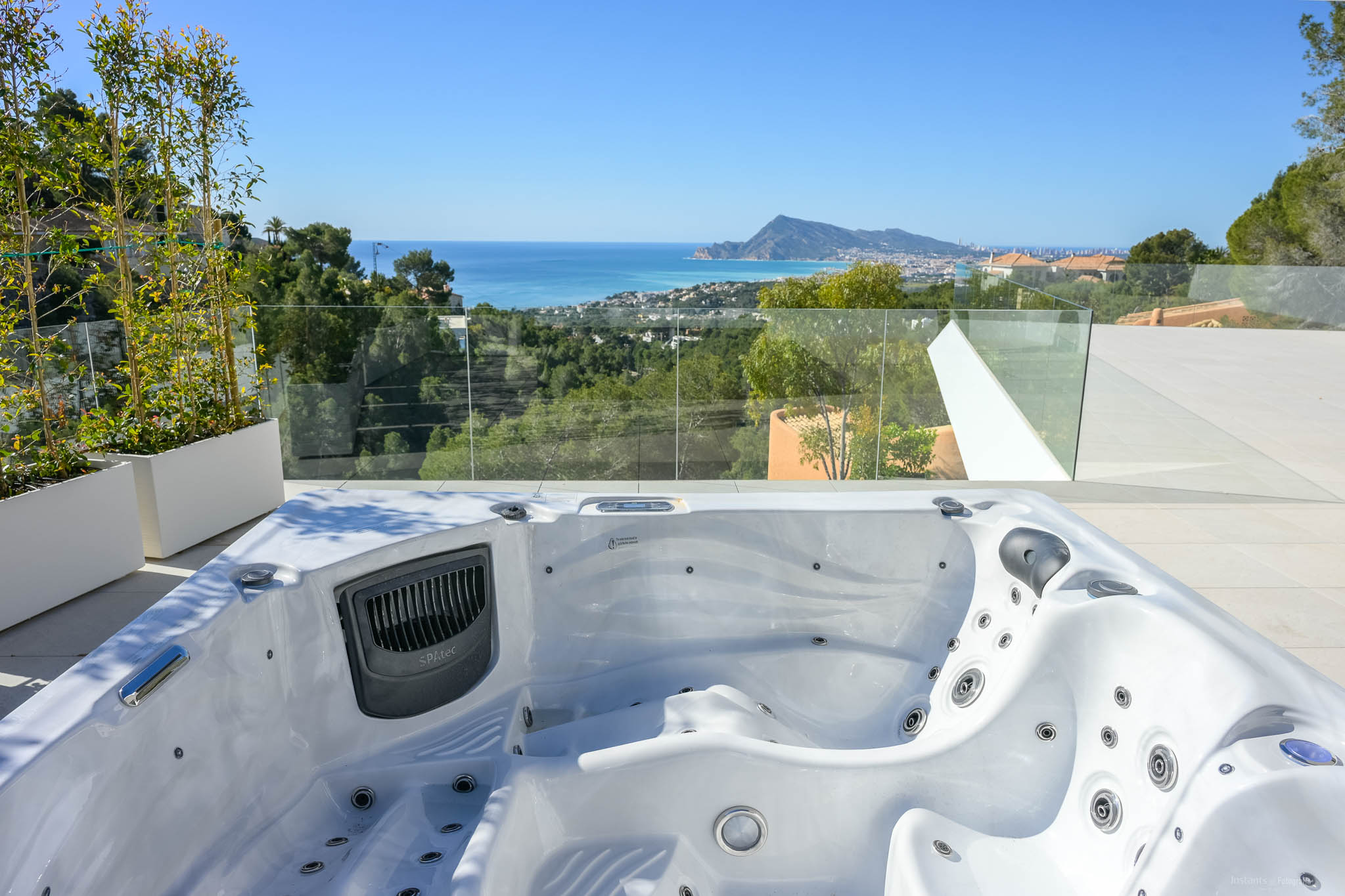 Außen-Whirlpool auf gefliester Terrasse mit Panoramablick auf Mittelmeer, Küstenstadt und Berge. Glasgeländer und Pflanzen akzentuieren die Aussicht.
