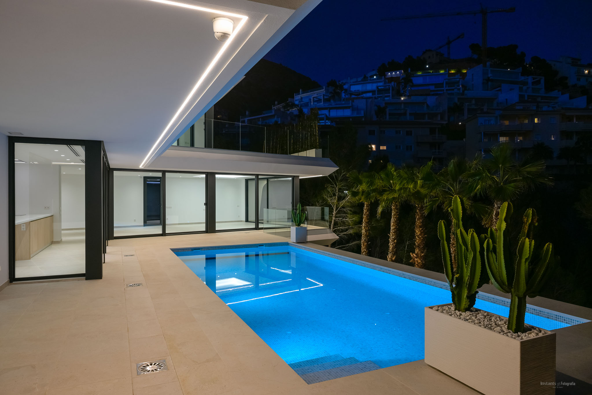 Beleuchteter Pool bei Nacht mit blauem Unterwasserlicht und gefliester Terrasse. Moderne lineare Deckenleuchten betonen die Architektur, mit Blick auf einen Hügel und entfernte Gebäude.
