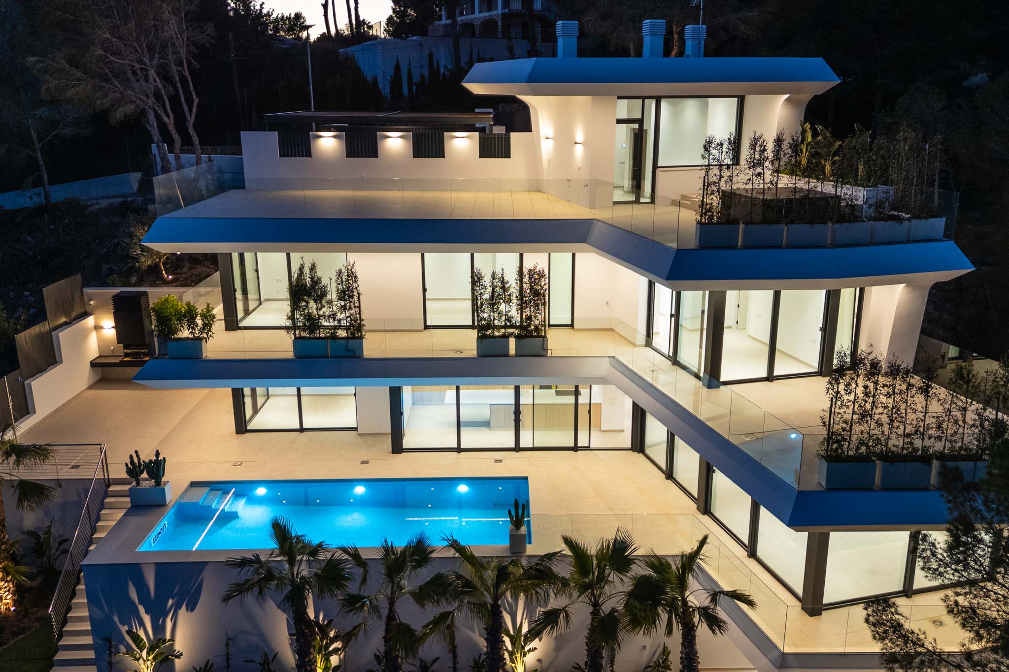 Die mehrstöckige moderne Architektur der Villa erstrahlt in der Dämmerung, mit einem leuchtend blauen Pool und weitläufigen Terrassen mit Glasgeländern.
