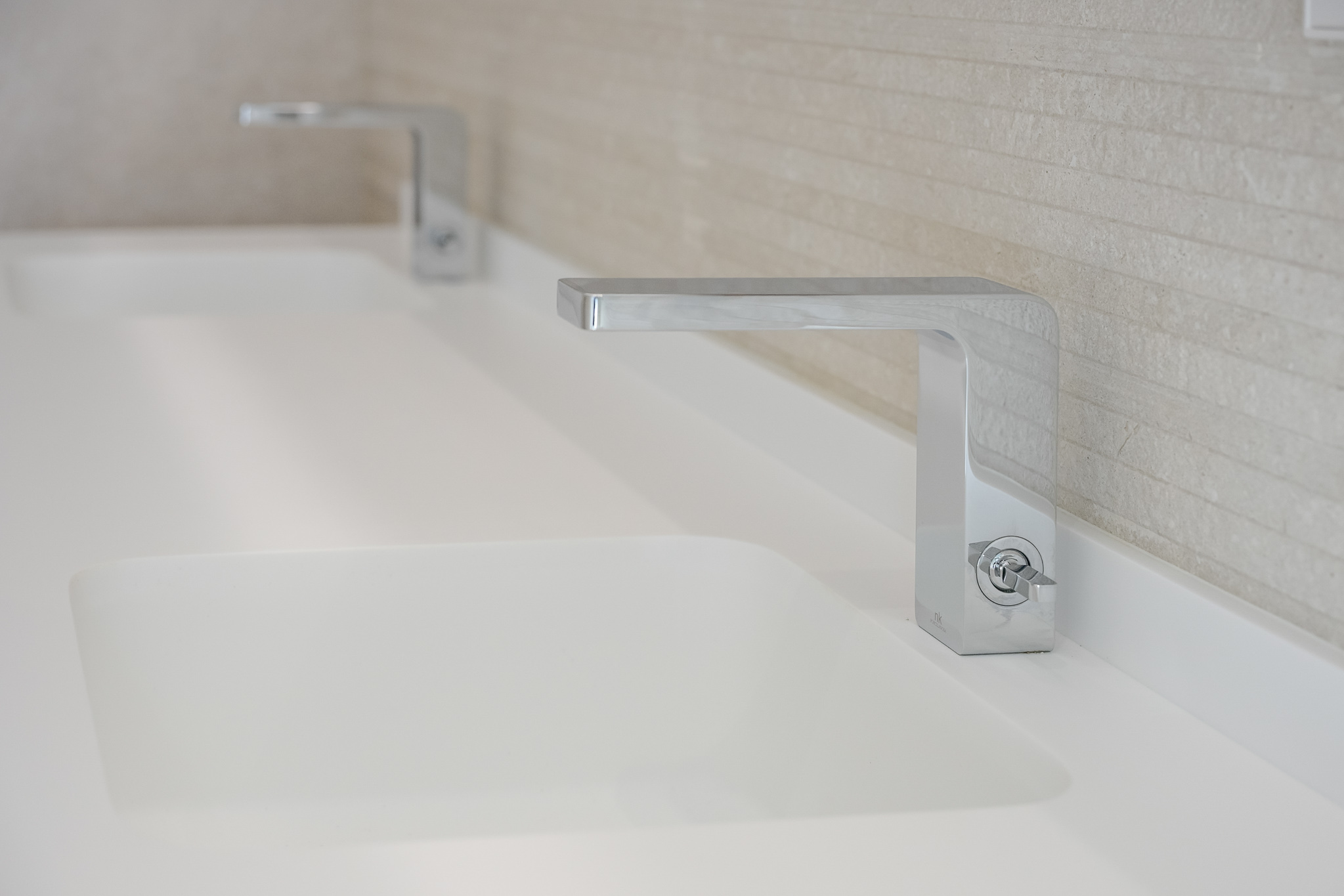 Doppelwaschtisch mit eleganten Chromarmaturen und integrierten weißen Waschbecken. Die Rückwand ist mit hellen, horizontal strukturierten Fliesen verkleidet.