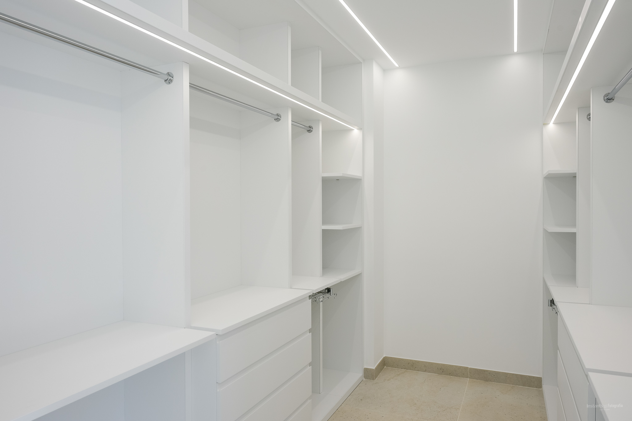 Geräumiger begehbarer Kleiderschrank mit maßgefertigten weißen Schränken, verchromten Kleiderstangen und integrierter LED-Beleuchtung. Bietet mehrere Regale und Schubladen auf hellem Fliesenboden.