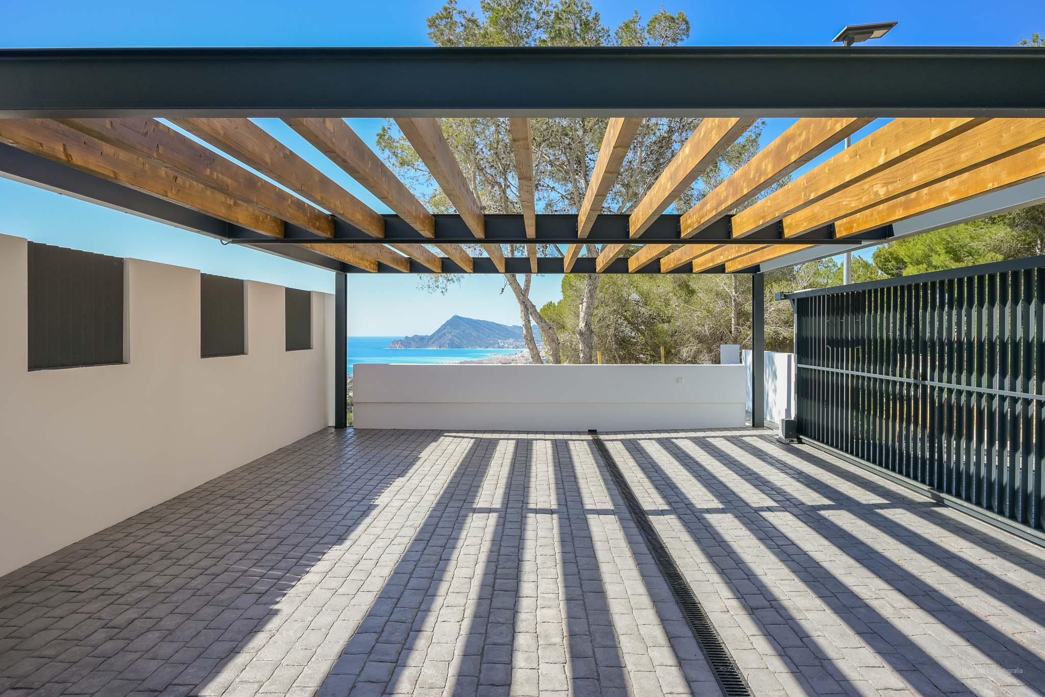 Großzügiger Außenbereich mit Pergola aus Holzlatten und Metallrahmen auf Kopfsteinpflaster. Bietet einen Panoramablick auf Meer und Berge, mit markanten Schatten durch das natürliche Licht.