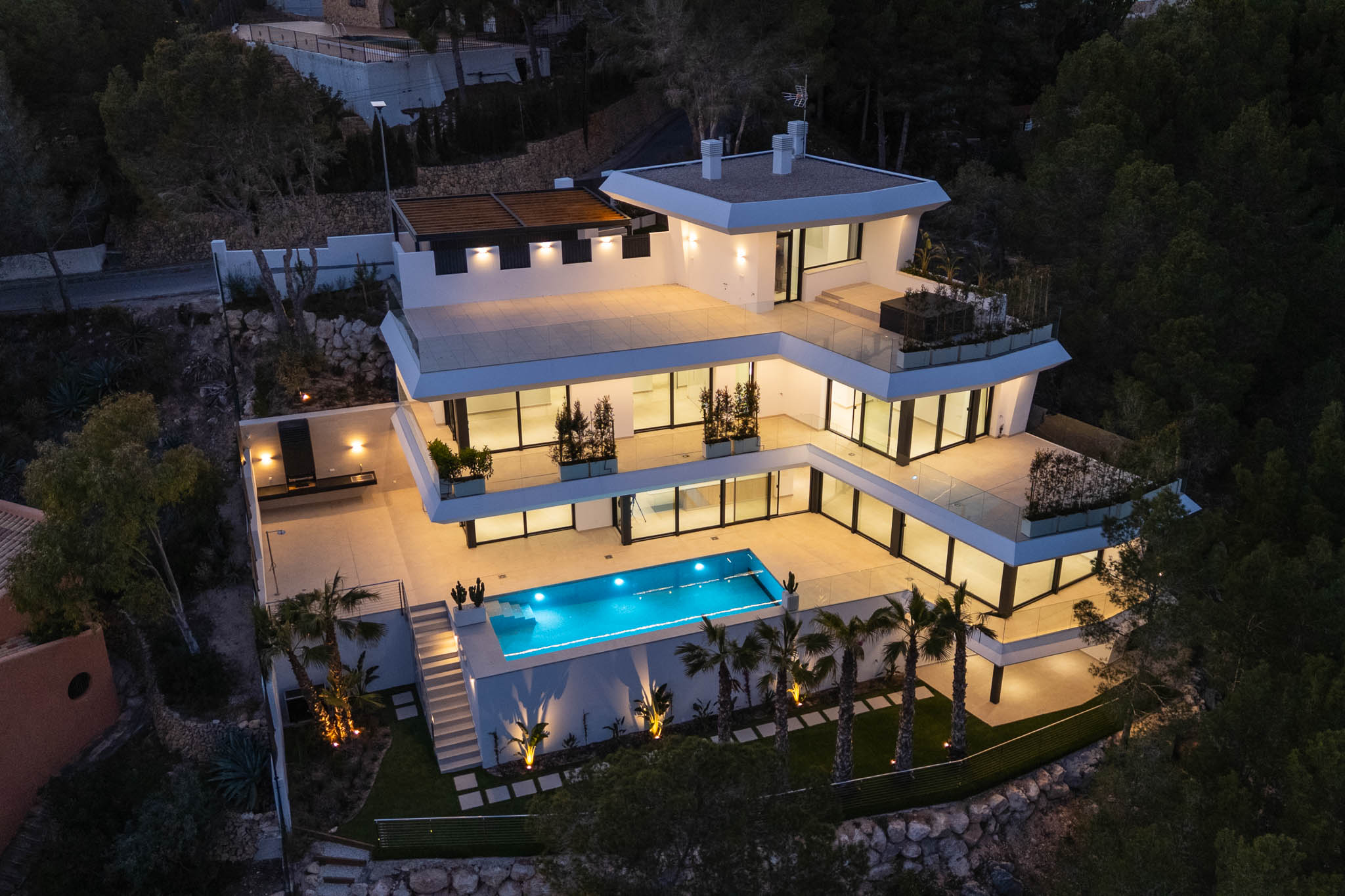 Luftaufnahme der modernen, mehrstöckigen Villa in der Dämmerung, mit beleuchteten Terrassen, einem hellblauen Pool und einer Außenküche, elegant beleuchtet inmitten üppiger Vegetation.
