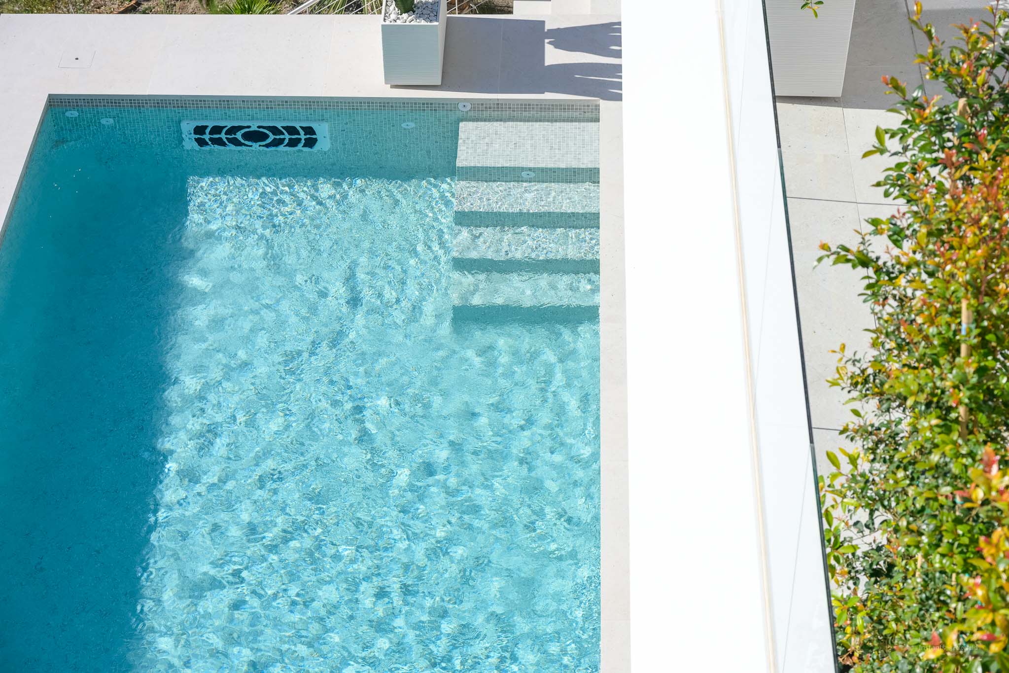 Luftaufnahme des hellblauen Mosaikpools mit integrierten Stufen, einem modernen Wasserrücklaufsystem und umliegendem hellem Pflaster.