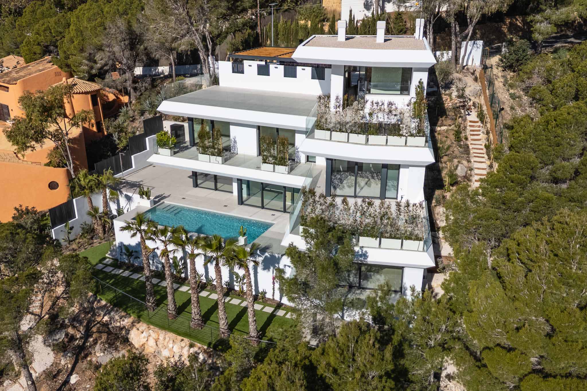 Luftaufnahme einer modernen, mehrstöckigen weißen Villa, eingebettet in einen grünen Hang. Mit großem privaten Pool, weitläufigen Terrassen mit Glasgeländer und Palmen, umgeben von mediterranem Wald.