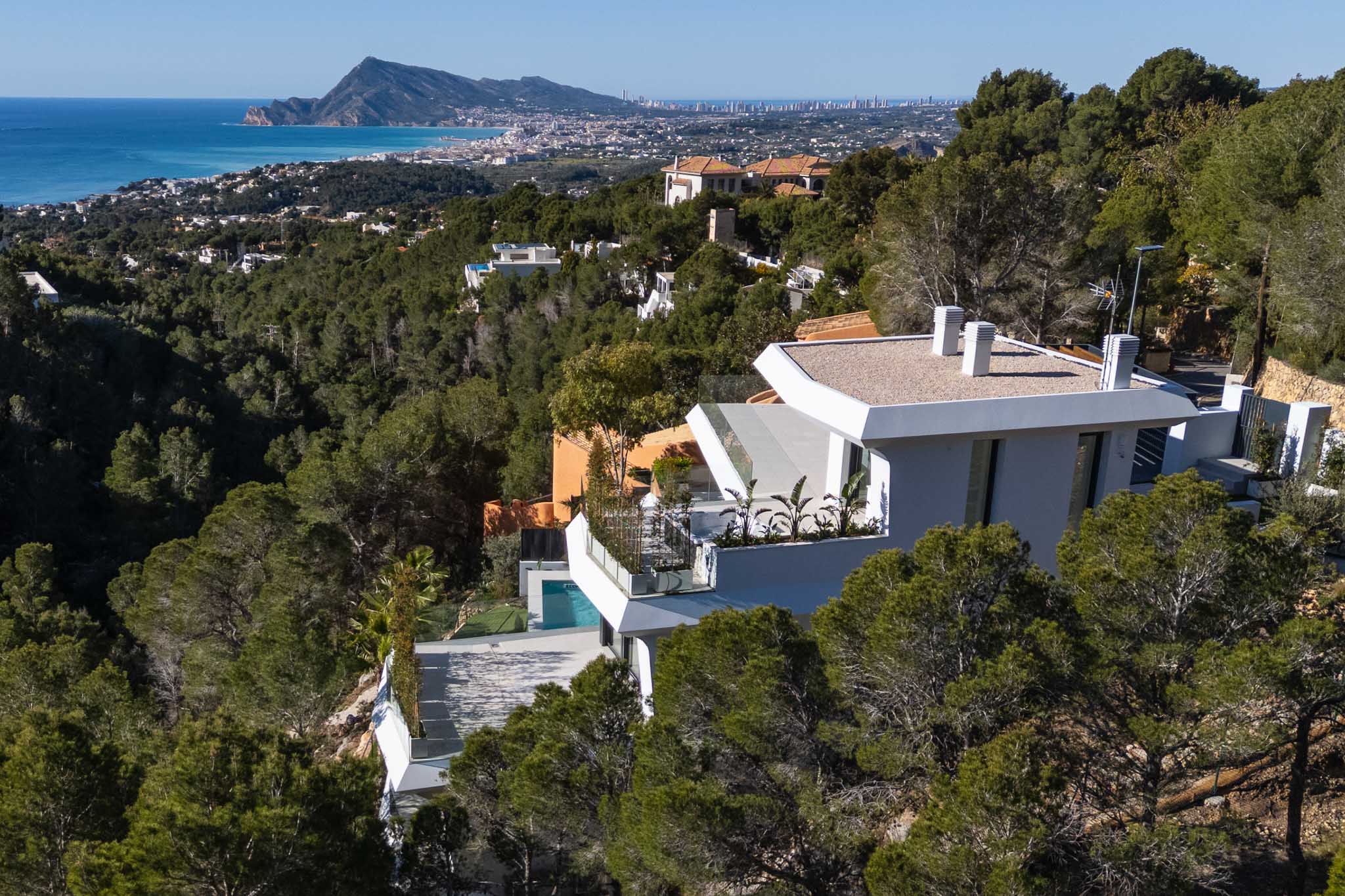Luftaufnahme einer modernen weißen Villa an einem pinienbewachsenen Hang, mit Panoramablick auf das Mittelmeer und die Küste von Altea. Ein privater Pool ist auf einer unteren Terrasse sichtbar.