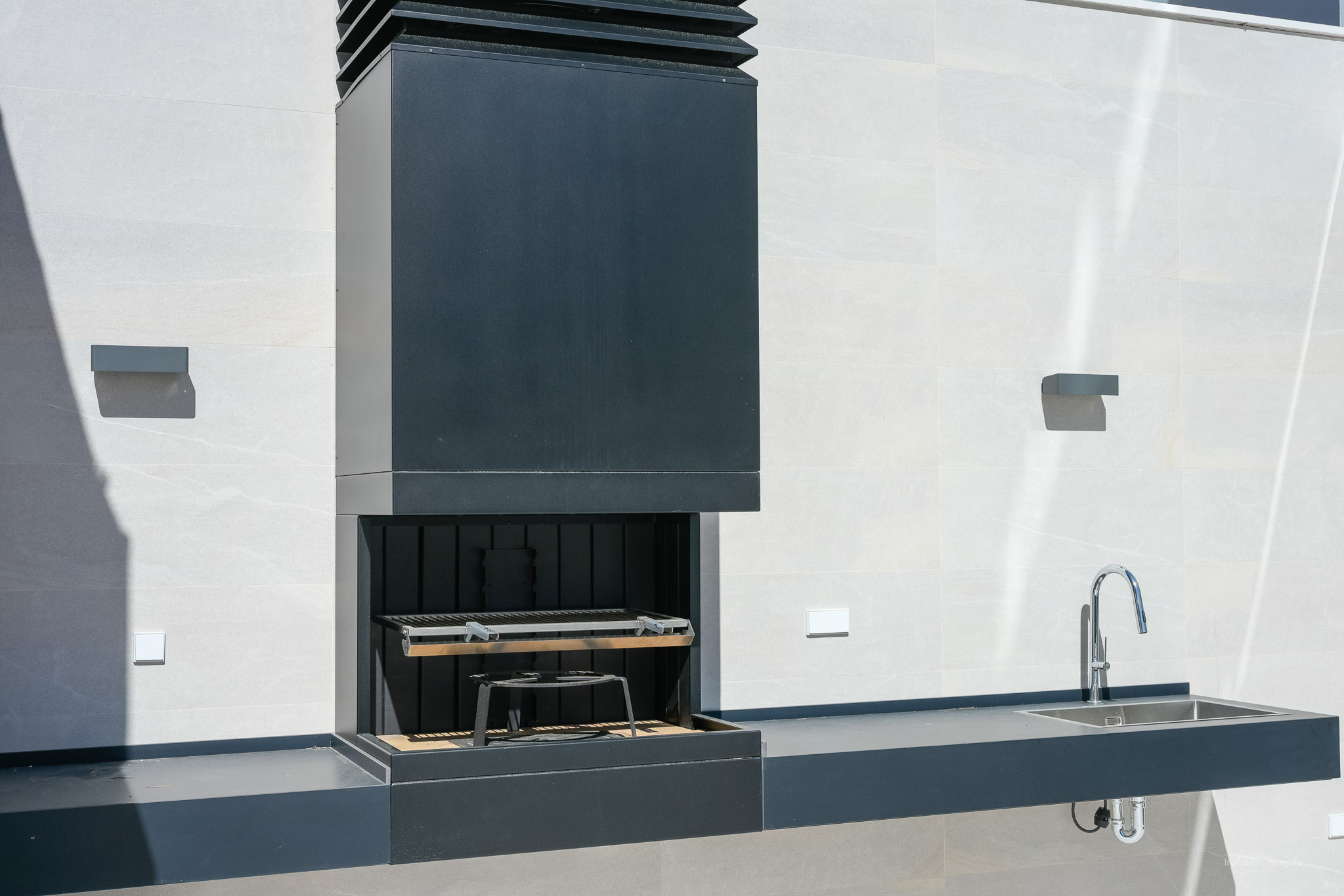 Moderne Außenküche mit dunklem Einbaugrill und Abzugshaube, passender Arbeitsplatte mit Edelstahlspüle und Chromarmatur, vor einer hell gefliesten Wand.