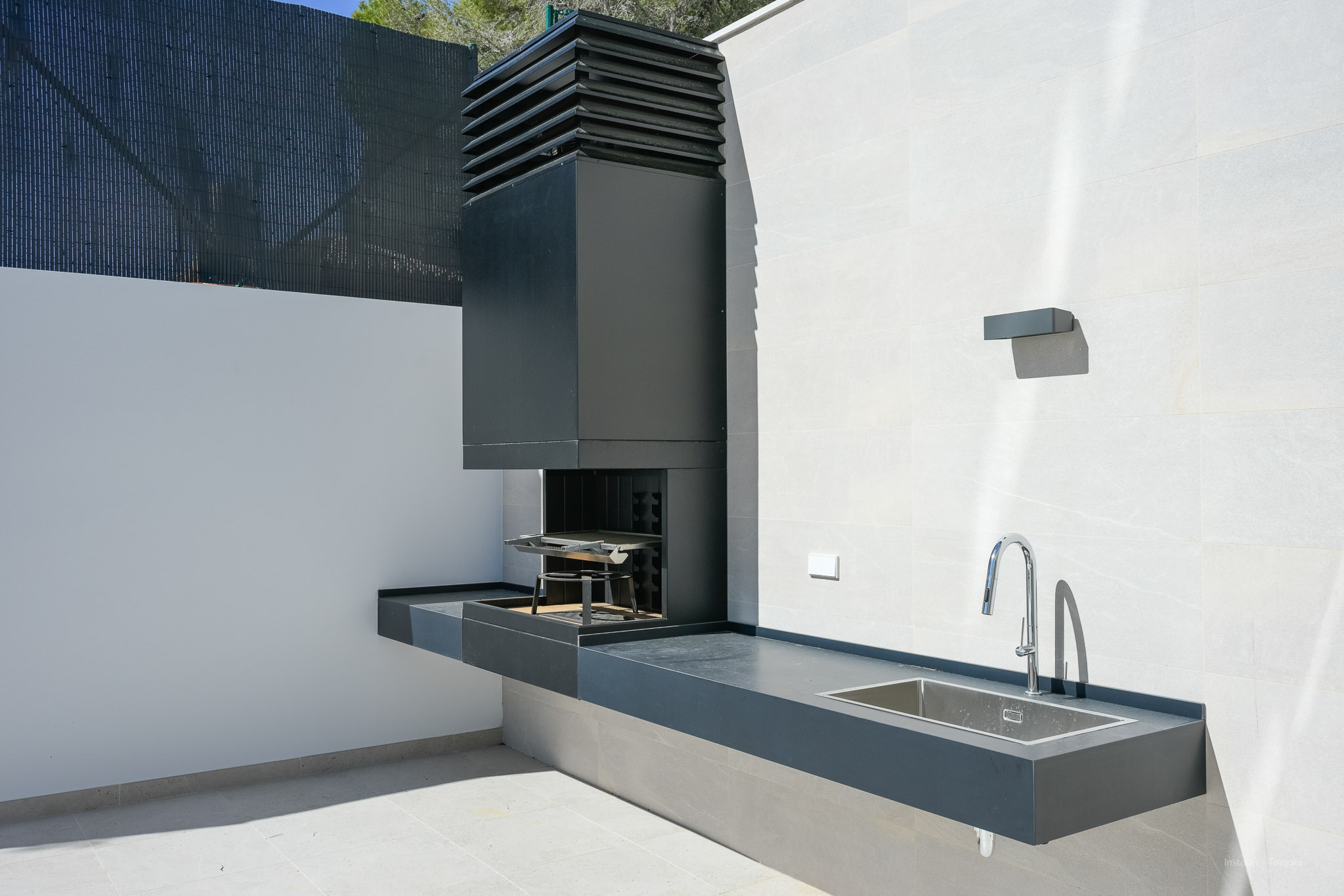 Moderne Außenküche mit integriertem dunkelgrauem Grill, passender Arbeitsplatte und Edelstahlspüle. Eine Chromarmatur ergänzt die helle Fliesenwand und den Boden.
