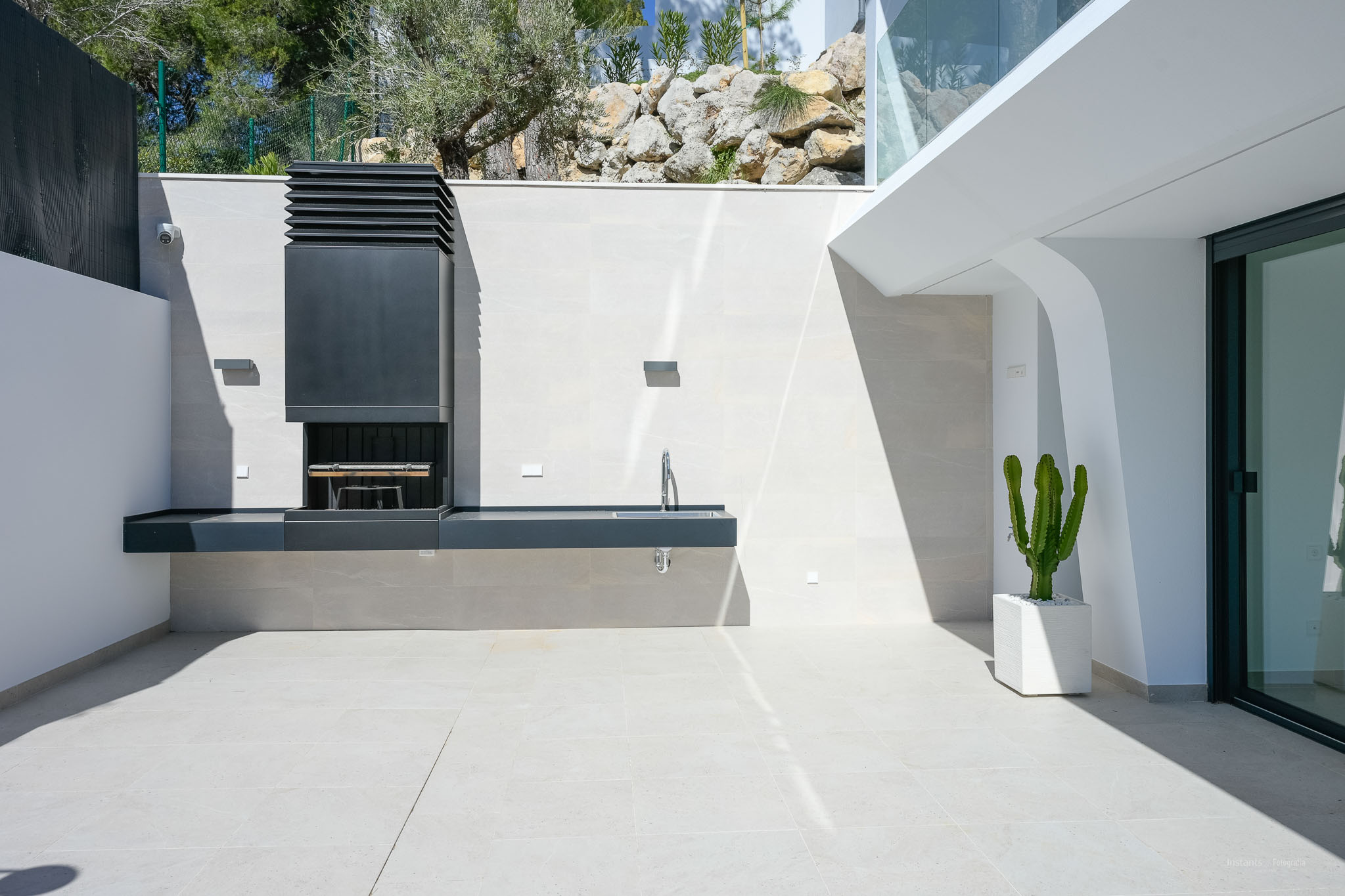 Moderne Außenküche mit integriertem Grill und Spüle auf hellen Fliesen. Ein Topfkaktus und natürliche Felslandschaft ergänzen den eleganten Außenbereich.