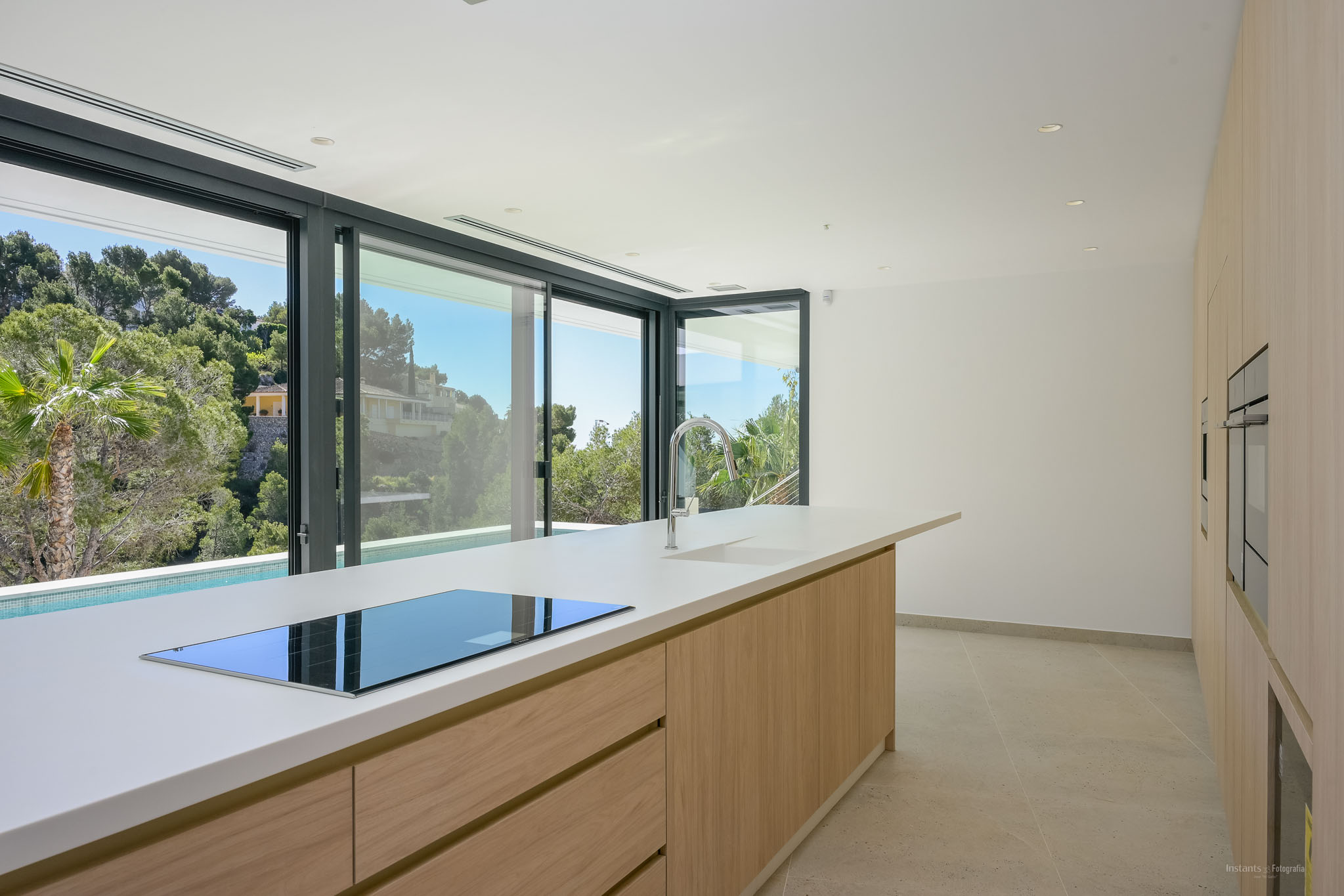 Moderne Küche mit großer Insel, weißer Arbeitsplatte, integriertem Induktionskochfeld und Chromarmatur. Schiebetüren bieten Blick auf mediterrane Vegetation und einen privaten Pool.