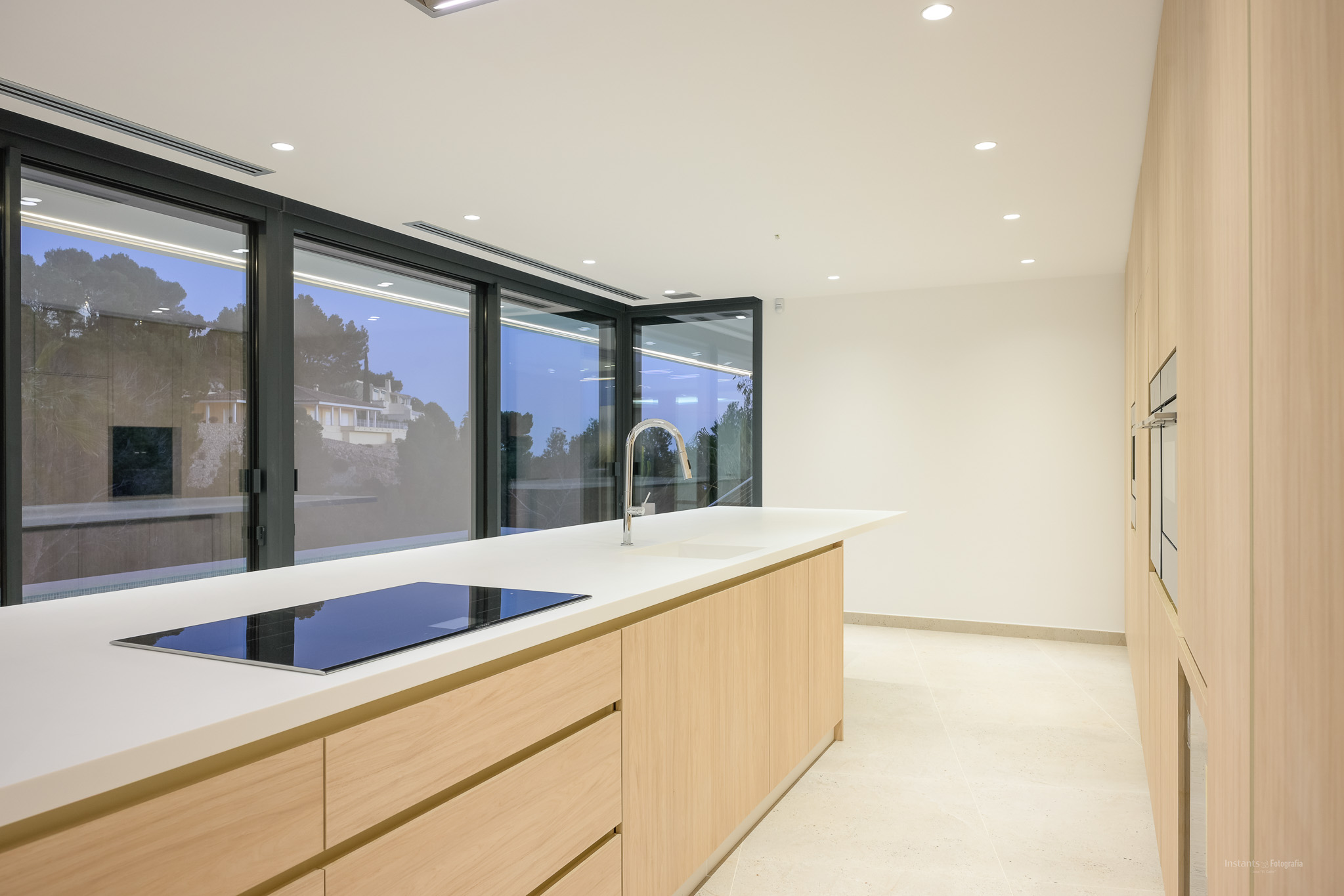 Moderne Küche mit großer Mittelinsel, weißer Arbeitsplatte und integriertem Induktionskochfeld. Helle Holzschränke und Einbauöfen an der Wand, ergänzt durch raumhohe Fenster.