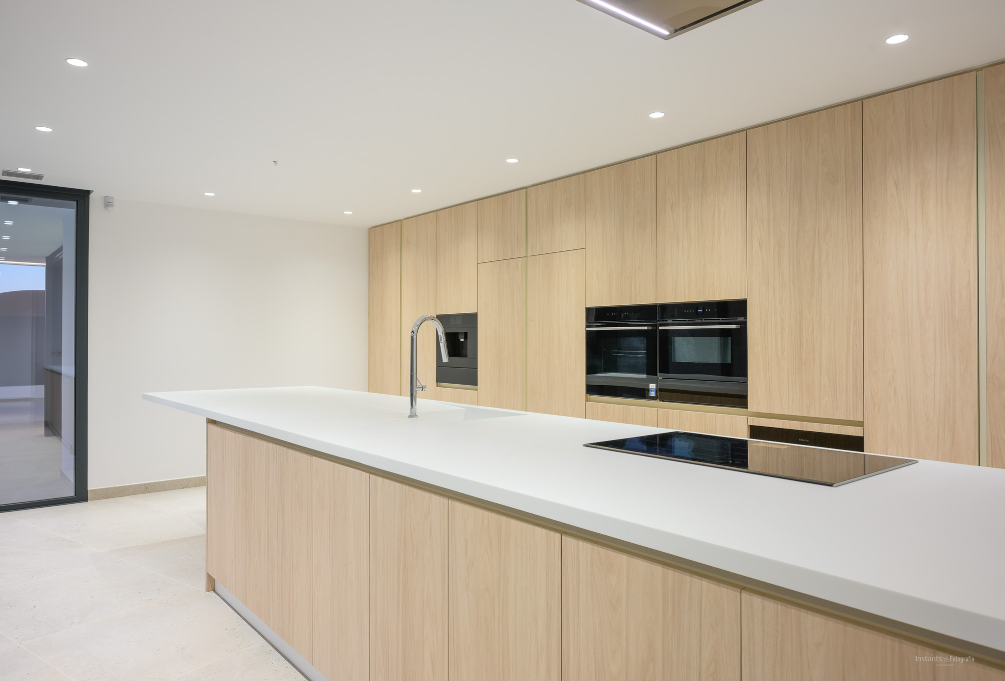 Moderne Küche mit großer weißer Insel, integrierter Spüle und Induktionskochfeld, ergänzt durch raumhohe helle Holzschränke und Einbaubacköfen.