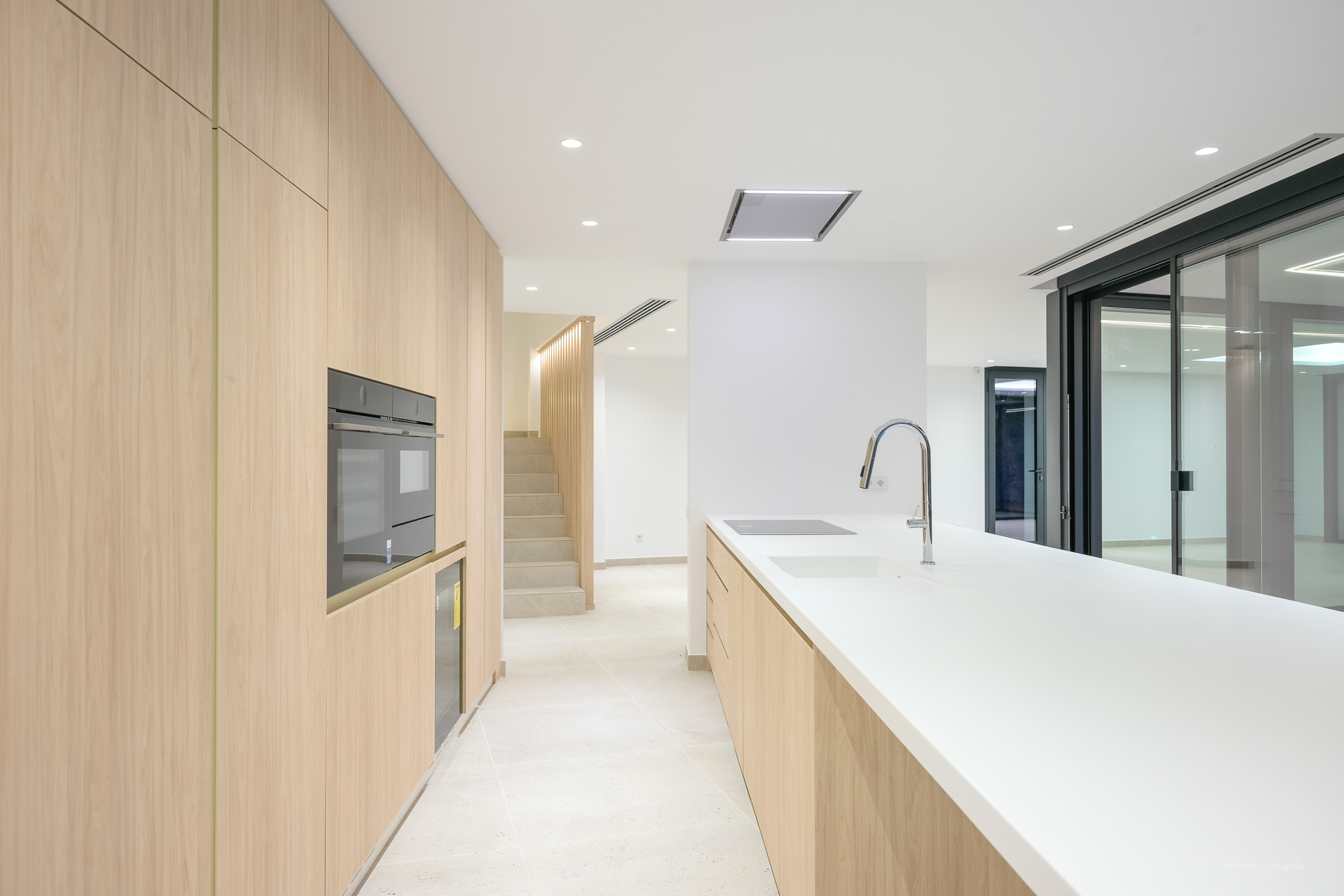 Moderne Küche mit hellen Holzschränken, weißer Arbeitsplatte und integrierten Geräten wie Backofen, Induktionskochfeld und Spüle mit Chromarmatur. Verbindung zu Treppe und Außenzugang.