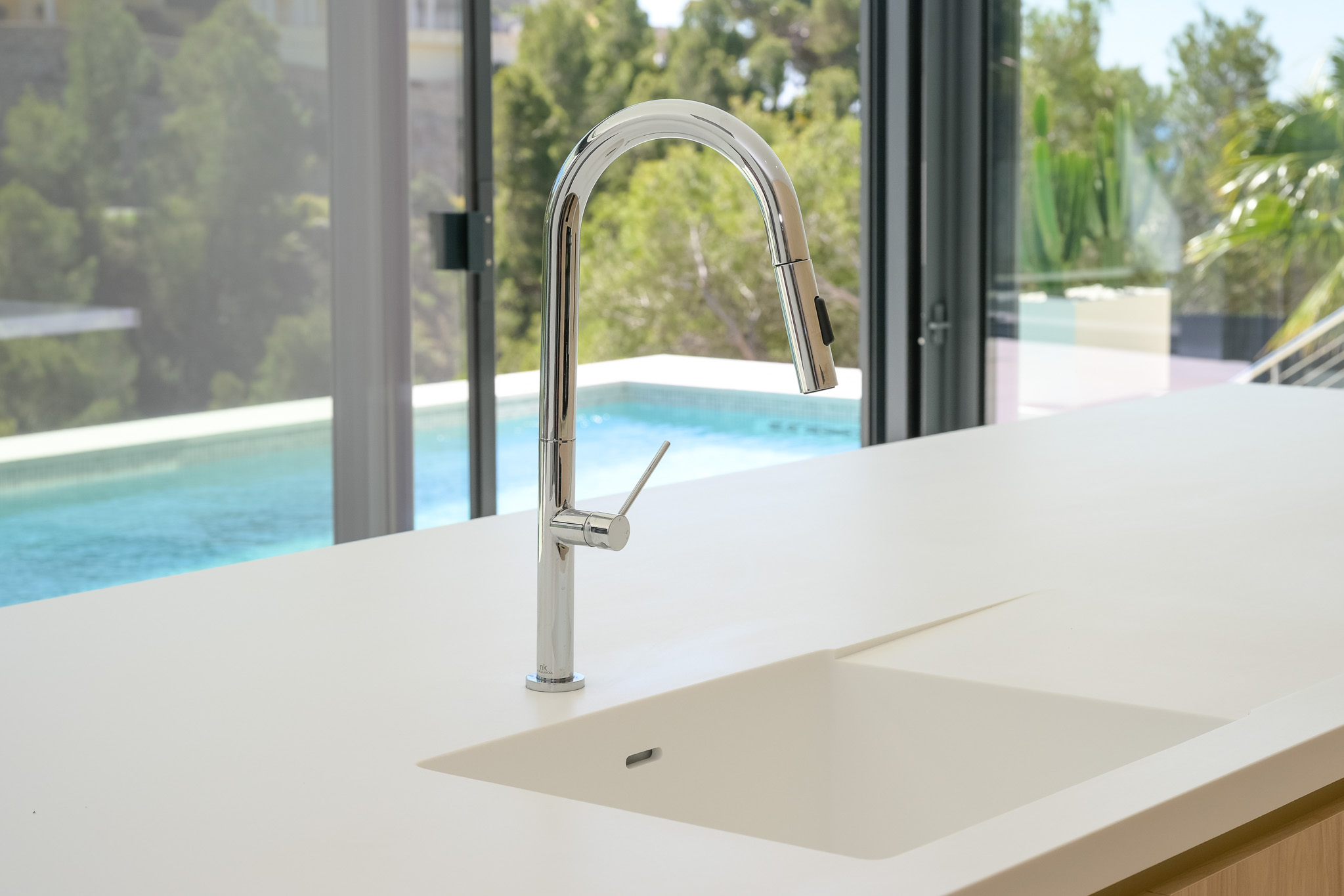 Moderne Küche mit heller Arbeitsplatte und integriertem Spülbecken. Ein eleganter Chrom-Ausziehhahn blickt auf den Pool und die grüne Außenanlage durch große Glasfronten.
