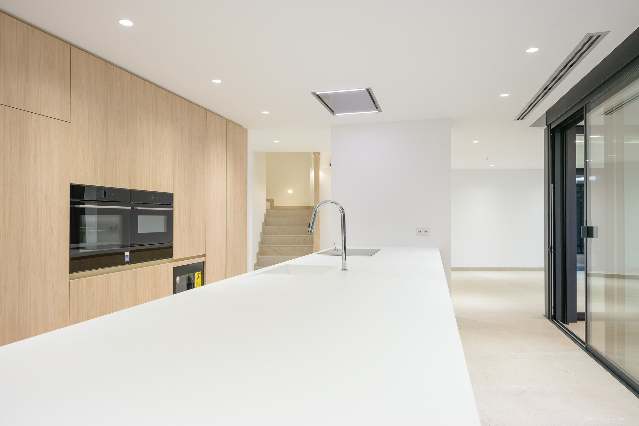 Moderne Küche mit langer weißer Insel, integrierter Spüle und Chromarmatur, ergänzt durch helle Holzschränke bis zur Decke und integrierte Doppelöfen. Einbauleuchten erhellen den zeitgenössischen Raum.