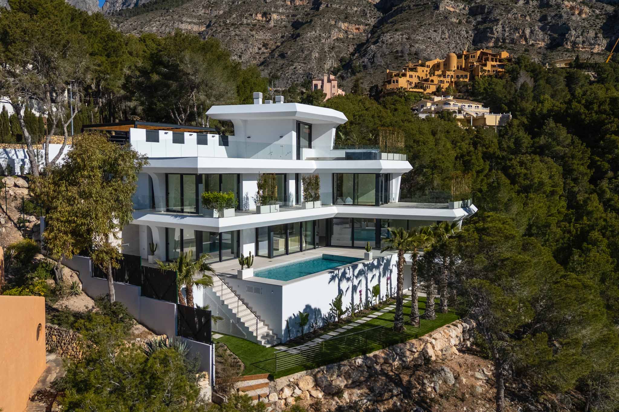Moderne, mehrstöckige Villa mit integriertem Rechteckpool und weitläufigen Glastterrassen, eingebettet in einen grünen Hang mit Bergblick.