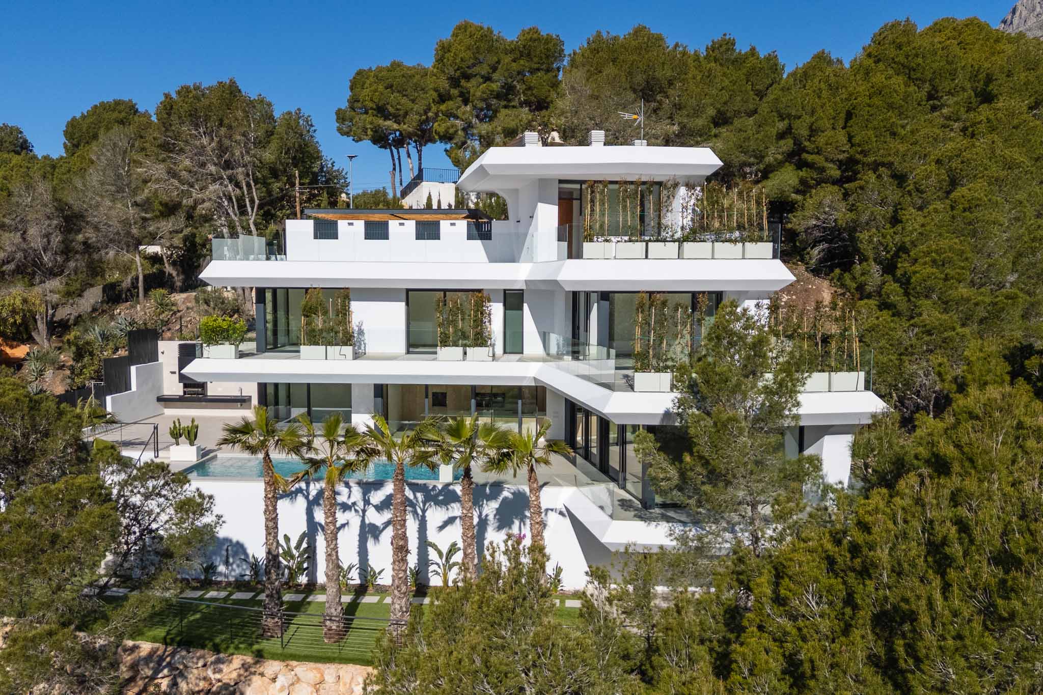 Moderne Villa mit mehreren Ebenen, weißer Fassade, großen Glast terrassen, einem Pool mit Palmen und Außenküche, eingebettet in eine grüne Hanglage.