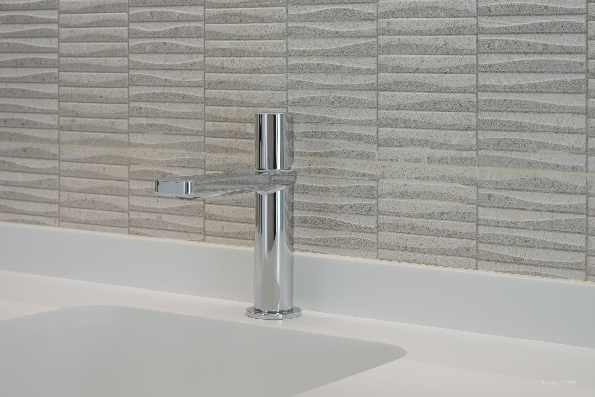 Moderner Chromwasserhahn auf einer weißen Arbeitsplatte mit integriertem Waschbecken. Die Wand ist mit strukturierten hellen Fliesen im Wellenmuster versehen.