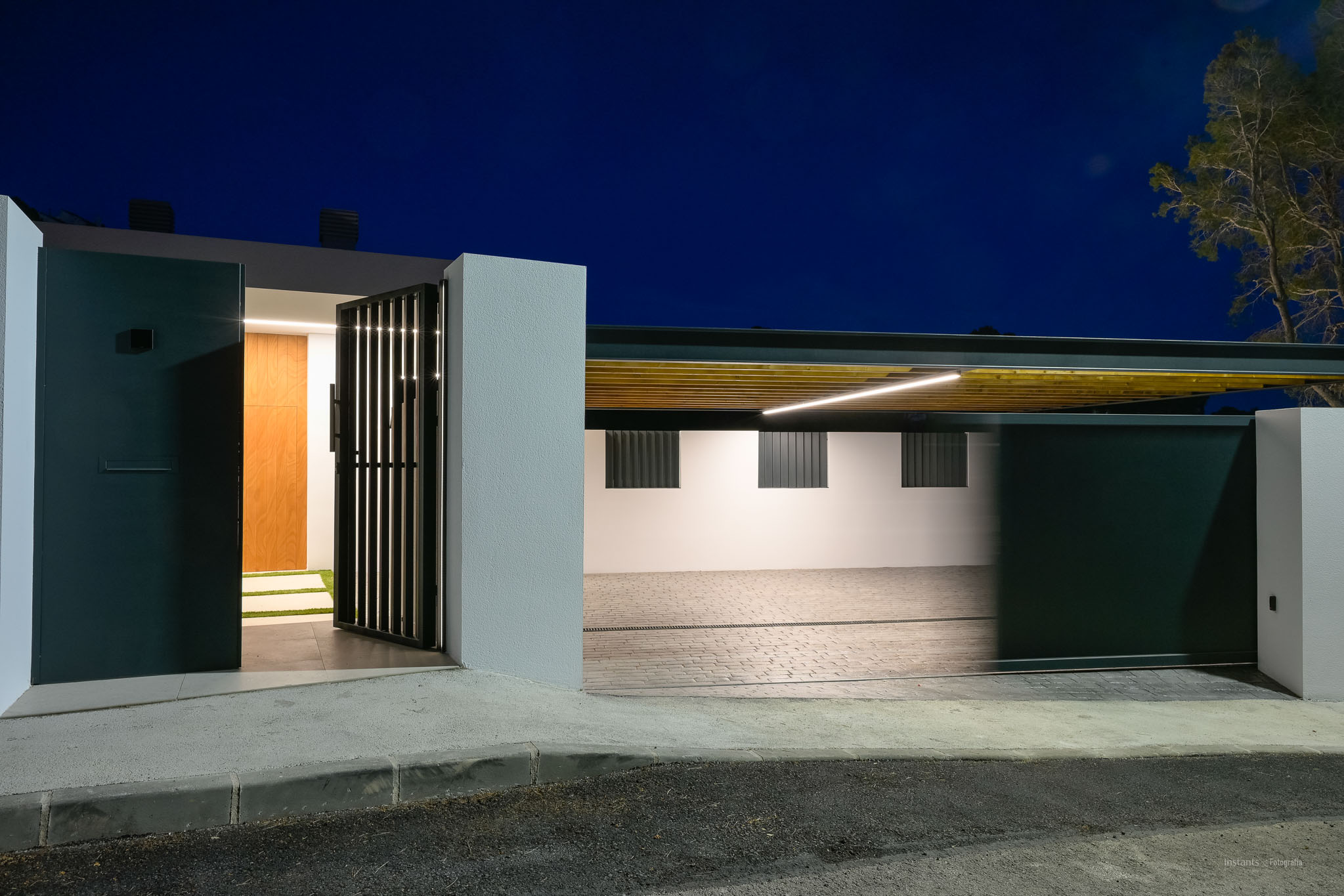 Moderner Eingangsbereich mit dunklem Fußgängertor und Holztür, daneben ein beleuchteter Carport mit Lamellendecke und linearer LED-Beleuchtung.