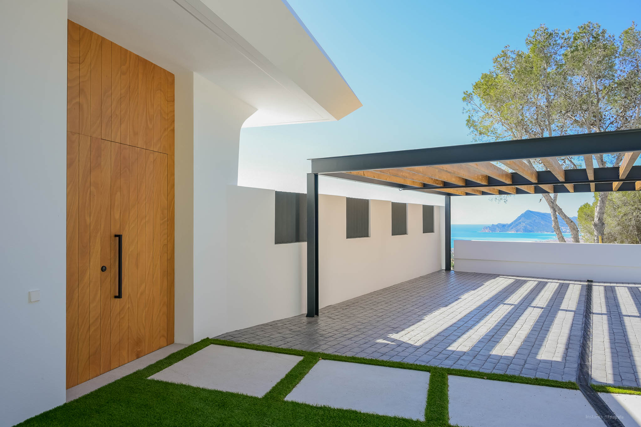 Moderner Eingangsbereich mit hoher Holztür, Kunstrasenweg und gepflastertem Parkplatz unter einer Metallpergola. Im Hintergrund sind das Mittelmeer und Berge sichtbar.