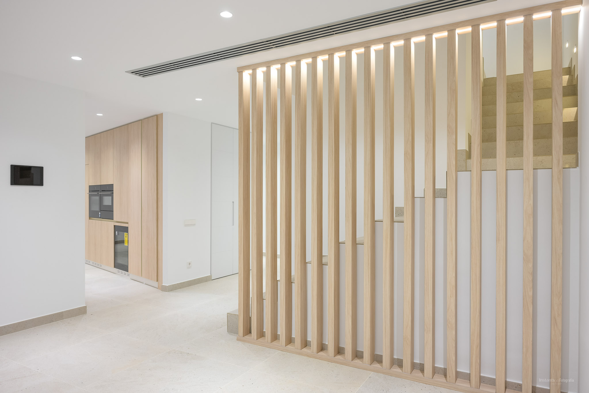 Moderner Innenraum mit heller Holzlamellenwand und integrierter Beleuchtung, die einen eleganten Küchenbereich mit Einbaugeräten von einer Steintreppe trennt.