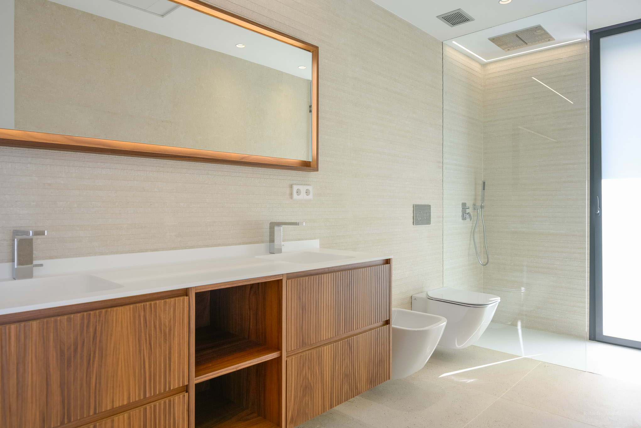 Modernes Badezimmer mit Doppelwaschtisch, Holzunterschränken und strukturierten Wandfliesen. Beleuchteter Spiegel, bodengleiche Dusche, wandhängendes WC und Bidet.