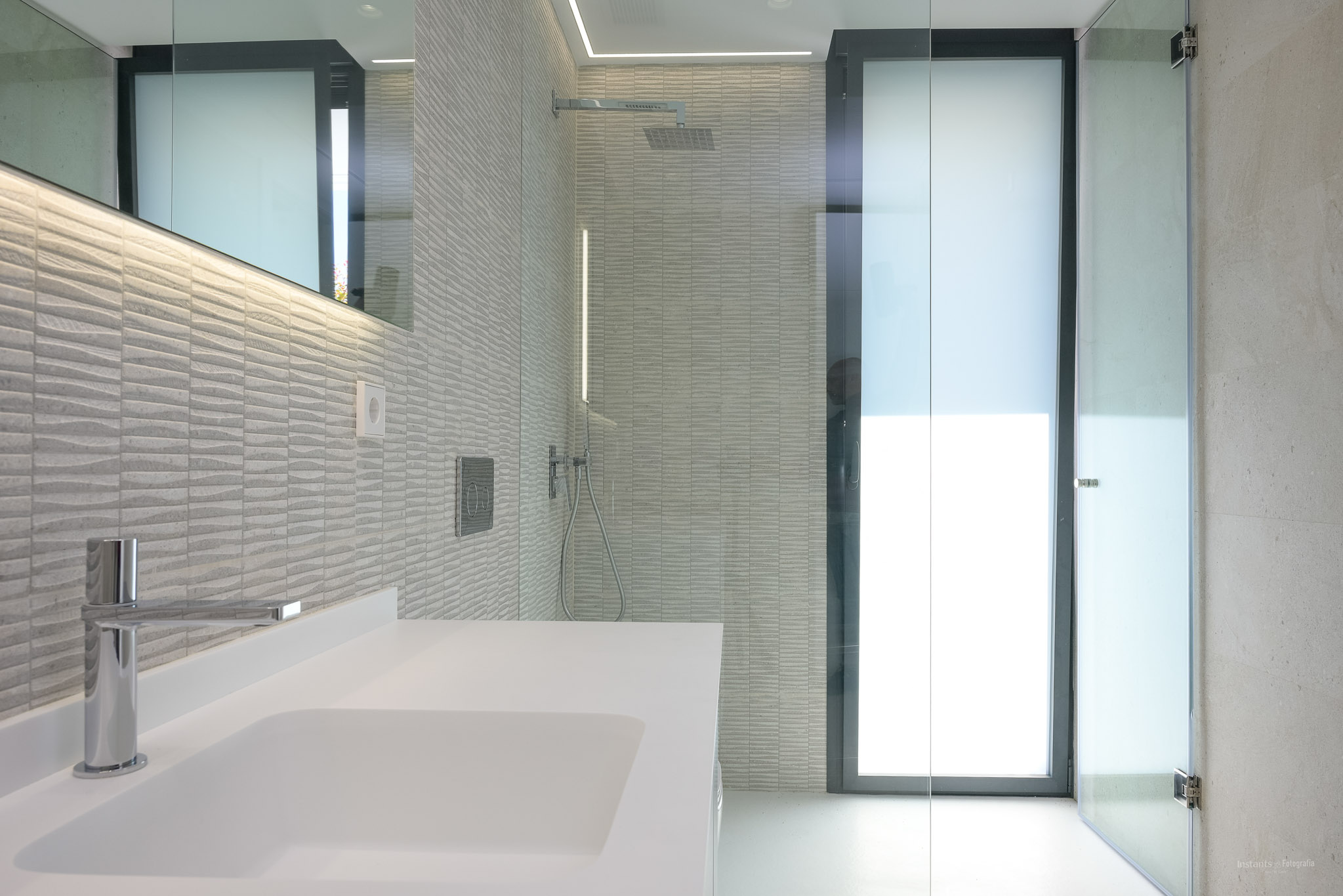 Modernes Badezimmer mit weißem Waschbecken, Chromarmatur und strukturierten grauen Wandfliesen. Es verfügt über eine Glasdusche mit Regenduschkopf und LED-beleuchteten Spiegel.