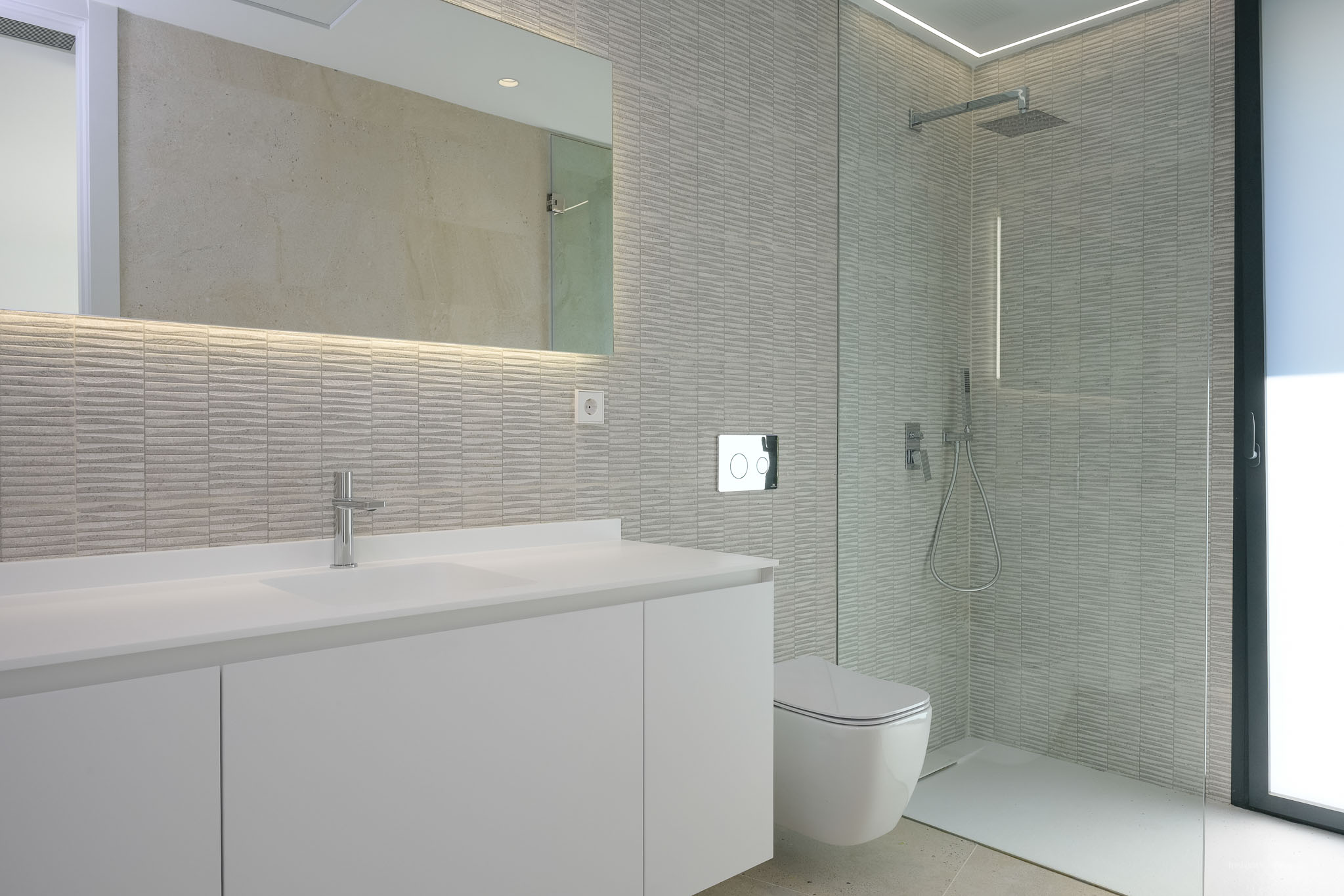 Modernes Badezimmer mit weißem Waschtisch, beleuchtetem Spiegel und strukturierten grauen Wandfliesen. Wandhängende Toilette und geräumige Duschkabine mit Glaswand.