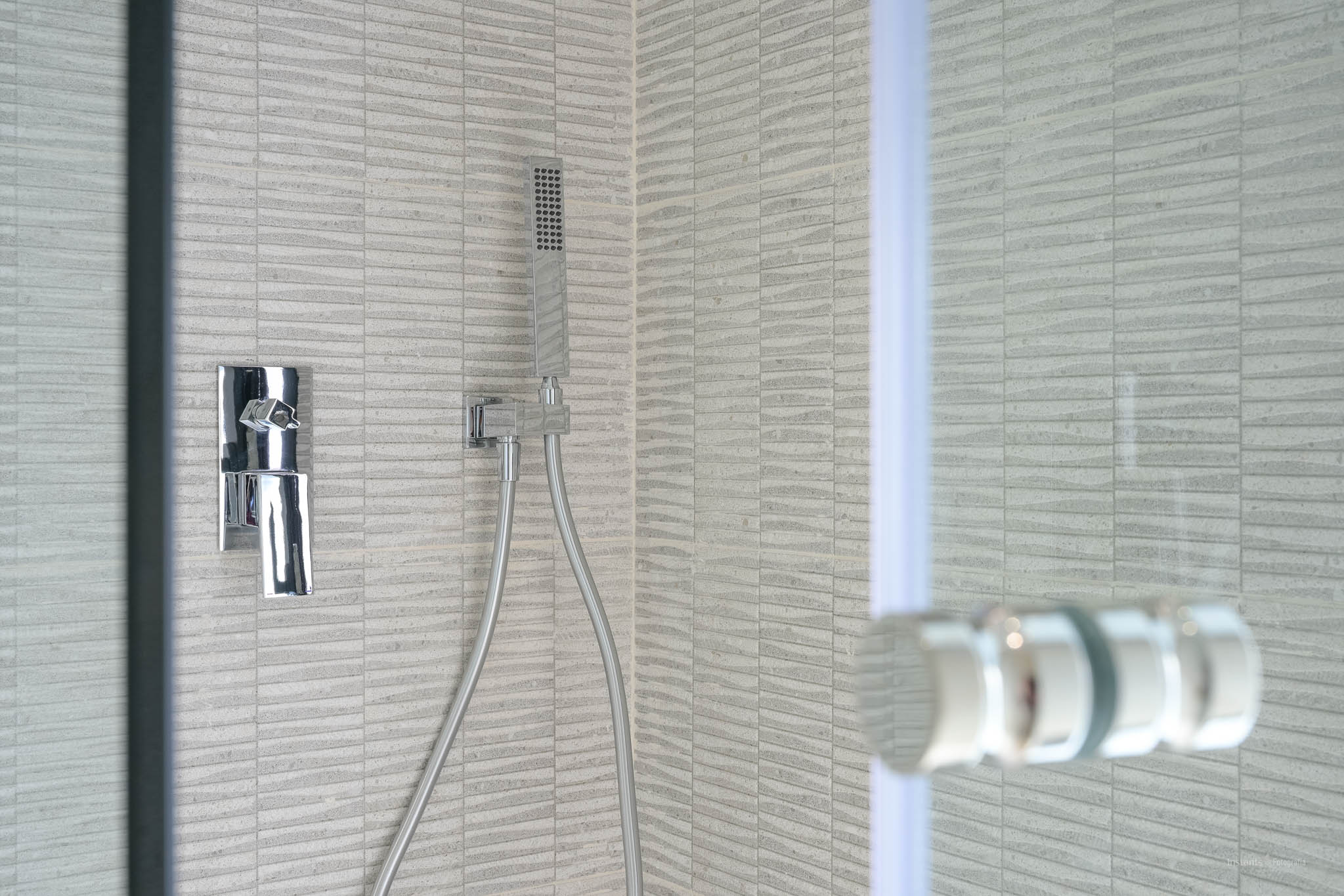 Modernes Duschdetail mit grauen, strukturierten Rechteckfliesen, einer eleganten Chrom-Mischbatterie und Handbrause. Eine Glasduschwand ist teilweise sichtbar.